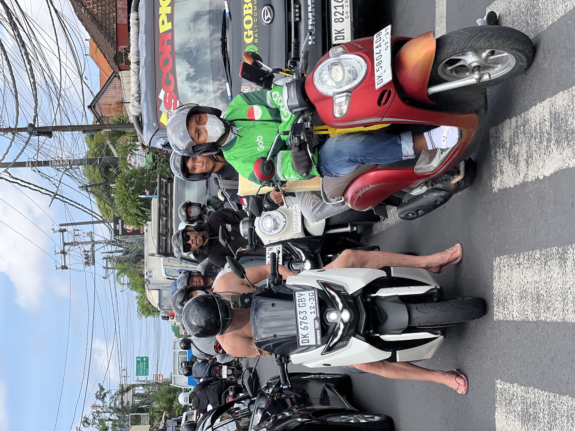 The image depicts a traffic jam in North Kuta, Indonesia. Multiple scooters and a black car are visible, all stationary in traffic. A man in the foreground, shirtless and wearing pink flip-flops, is on a white scooter with a license plate reading "DK 6763 GBY". To his right, a person wearing a green jacket with a "Grab" logo and a face mask is on a red scooter with a license plate reading "DK 5804 AD8". The background shows buildings with tiled roofs and a sky with some clouds. Overhead, numerous electrical wires crisscross the scene, indicative of urban infrastructure. The road has a pedestrian crossing in the foreground.