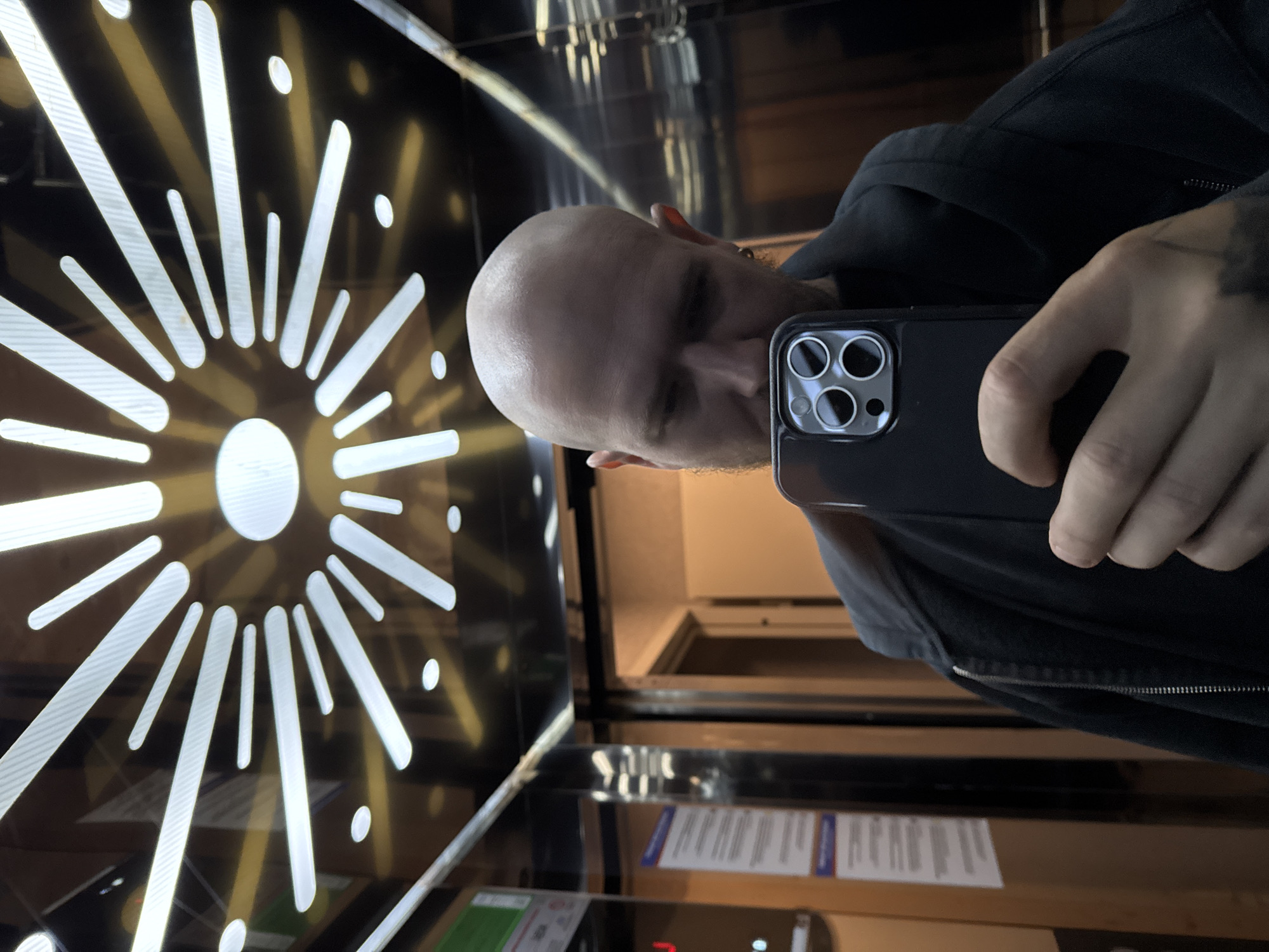 A bald man is taking a selfie in what appears to be an elevator. He is holding a black iPhone in his right hand. The elevator walls are mirrored with a geometric design featuring white radiating lines and circles. The floor selection panel is visible at the bottom of the frame. The location is Arnavutköy, Turkey, as indicated by the elevator's interior design, which is characteristic of buildings in that city. The man is wearing a dark-colored hooded jacket.
