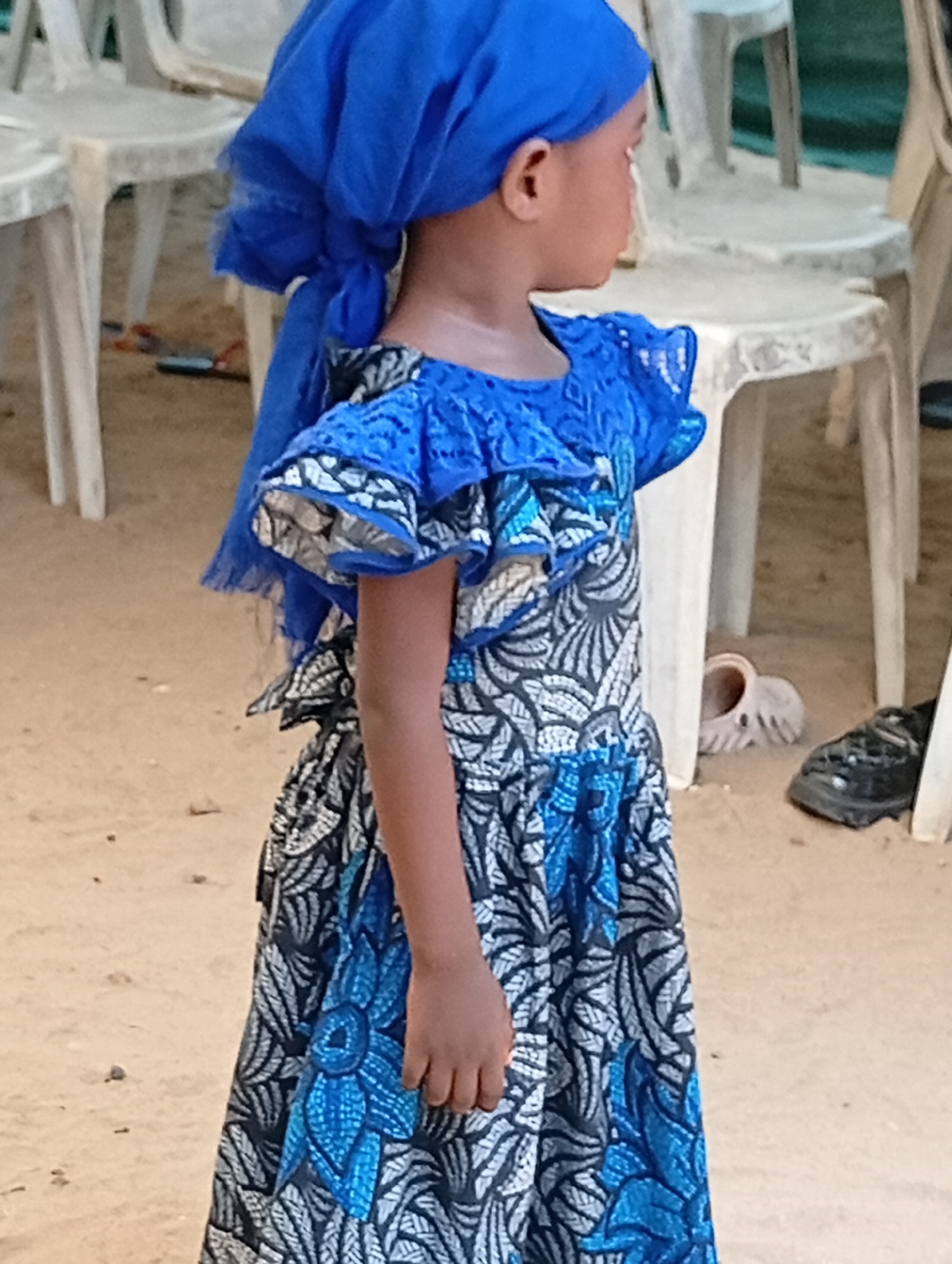 A young child stands in Adankolo, Nigeria, wearing a blue headscarf and a patterned dress with blue accents. The background consists of sandy ground and white plastic chairs. A pair of shoes are visible under one of the chairs.