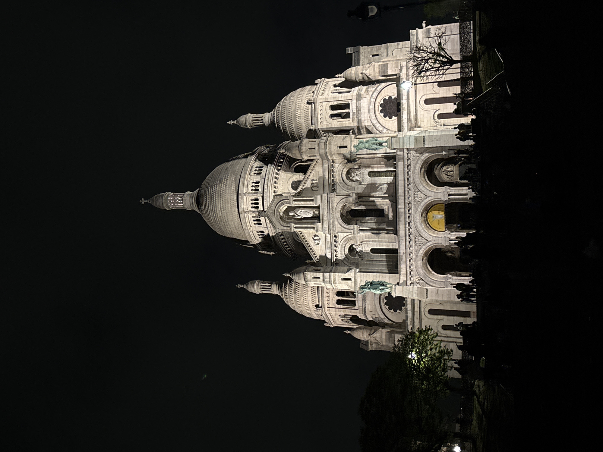 The image is a night shot of the Sacré-Cœur Basilica in Paris, France. The basilica is illuminated and appears as a large, light-colored structure against a dark sky and foreground. The architectural details, including the domes, arches, and statues, are visible. A few trees and a lamppost are seen in the foreground. Some silhouetted figures can be seen at the base of the building.