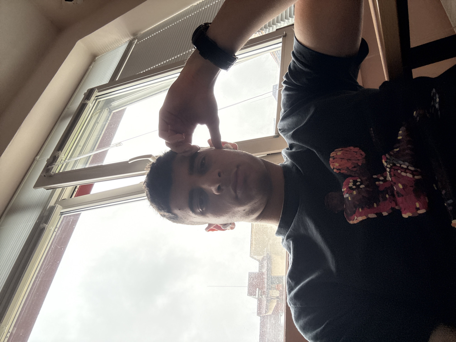 A young man is captured in a casual, low-angle selfie, with the image rotated 90 degrees clockwise from its likely original orientation. He is looking directly at the camera with a neutral expression. His right arm is raised, with his hand near his right temple or ear, possibly adjusting something or making a subtle gesture. A dark-colored smartwatch or fitness tracker is visible on his right wrist.
He is wearing a dark t-shirt featuring a prominent design on the chest composed of red, orange, and white speckled patterns, resembling abstract art or potentially stylized butterflies.
The background is largely dominated by a large window with white frames, parts of which appear to be slightly ajar. Through the window, an overcast sky is visible, suggesting cloudy weather during daylight hours. Above the window, some internal blinds or ventilation slats can be seen, and a section of a light-colored ceiling or wall is also visible in the upper part of the frame (when oriented correctly).
The scene is indoors, likely in a room in Sofia, Bulgaria, given the provided context. No visible text is present in the image.