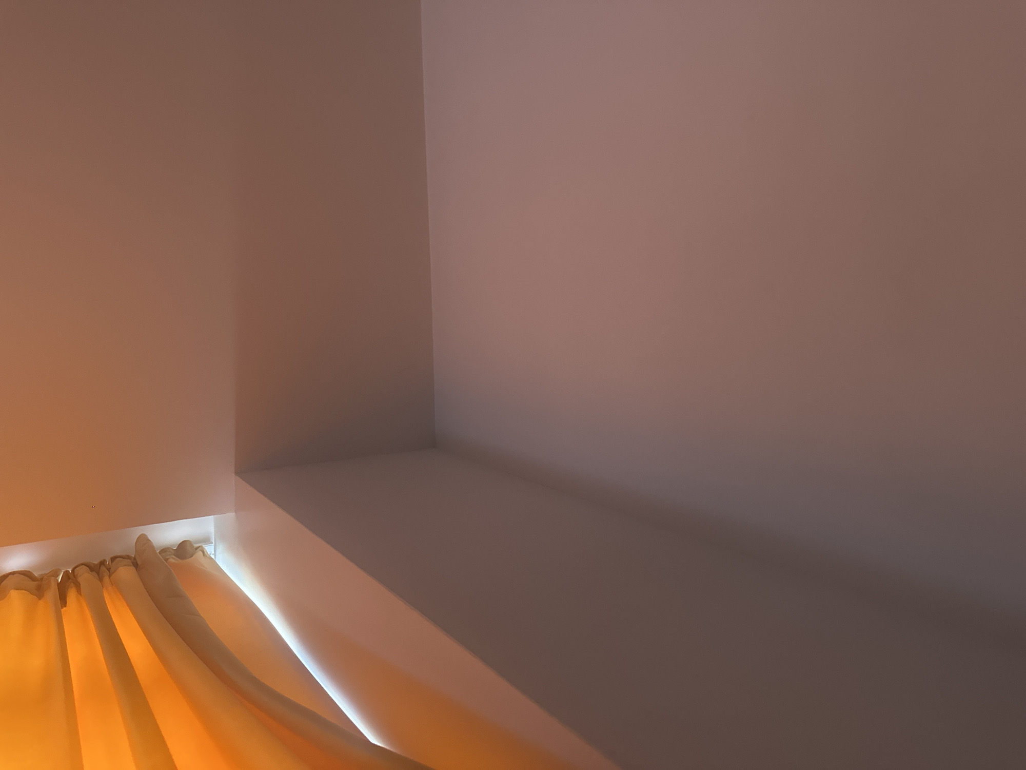 The image shows a close-up view of an interior corner where a wall meets a sloped ceiling, with orange curtains partially visible in the upper left. The lighting suggests it is daytime, with light filtering through the curtains and casting shadows. The walls and ceiling appear to be painted a light, neutral color. There are no people or other objects clearly visible in the scene.