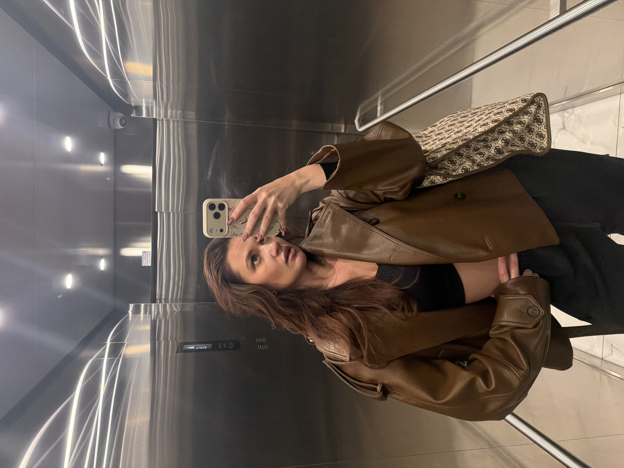 The original image, presented rotated 90 degrees clockwise, depicts a young woman taking a mirror selfie inside a modern elevator. She stands upright, reflected in the highly polished, metallic walls, which are likely stainless steel. The woman has long, wavy brown hair and is wearing a brown, possibly faux-leather, jacket over a black crop top and dark pants. Her left hand holds a light-colored smartphone with a subtle, possibly floral-patterned case, while her gaze is directed slightly upwards and to her right, away from the phone's screen. Her expression is neutral and contemplative.
To her right, a large patterned handbag with brown trim is visible. The pattern prominently features repeating "G" logos, indicating it is likely a Guess brand bag. The elevator's interior is illuminated by bright artificial lighting, including strip lights that create long, glowing reflections on the metallic surfaces and several recessed ceiling lights. A white security camera (CCTV) is mounted high on one of the metallic walls, and a metal handrail runs along another. The floor appears to be a light-colored tile, possibly marble. An elevator control panel with illuminated buttons is also reflected on the wall beside the woman. The scene captures a casual, indoor moment typical of an urban environment, potentially in Belgrade, Serbia.