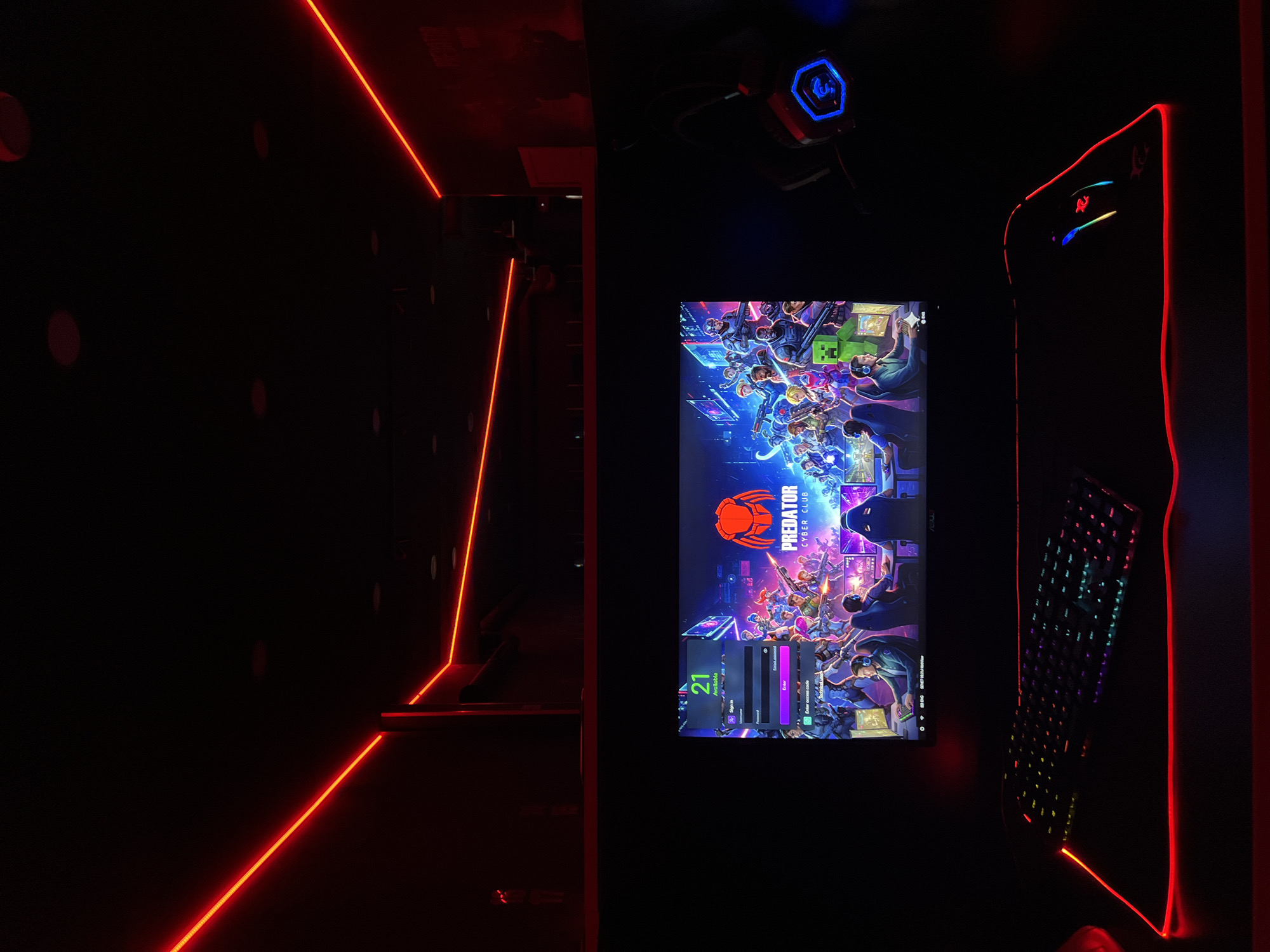 This image depicts a dark, futuristic-themed gaming setup within what appears to be a cyber cafe, located in Budva, Montenegro. The scene is illuminated primarily by artificial lights, creating a vibrant yet focused atmosphere.
A gaming monitor dominates the central area, displaying a login screen for "PREDATOR CYBER CLUB." The screen features a colorful, action-packed digital illustration with multiple animated characters, typical of online games or gaming platforms. Visible text on the login interface includes "21," "Anonimno," "Sign Up," "Enter access code," and "Enter."
To the right of the monitor, a gaming keyboard with dynamic, multi-colored RGB backlighting is present. A desktop computer tower, partially visible behind the keyboard, glows with red LED accents, contributing to the overall aesthetic. Above the monitor, a gaming headset, possibly from the "REDDRAGON" brand due to its logo and blue LED illumination on the earcup, hangs ready for use.
The room's walls are dark, punctuated by subtle, circular patterns or panels. Bright red LED strip lights delineate the edges of the walls and possibly the floor, forming crisp, geometric lines that guide the eye and add to the high-tech ambiance. These red lights are a significant source of illumination in the otherwise dim space.
The setup suggests an environment designed for immersive gaming, ready for activity, although no people are visible. The combination of deep shadows and vivid, colored lights conveys an energetic and modern gaming club atmosphere.