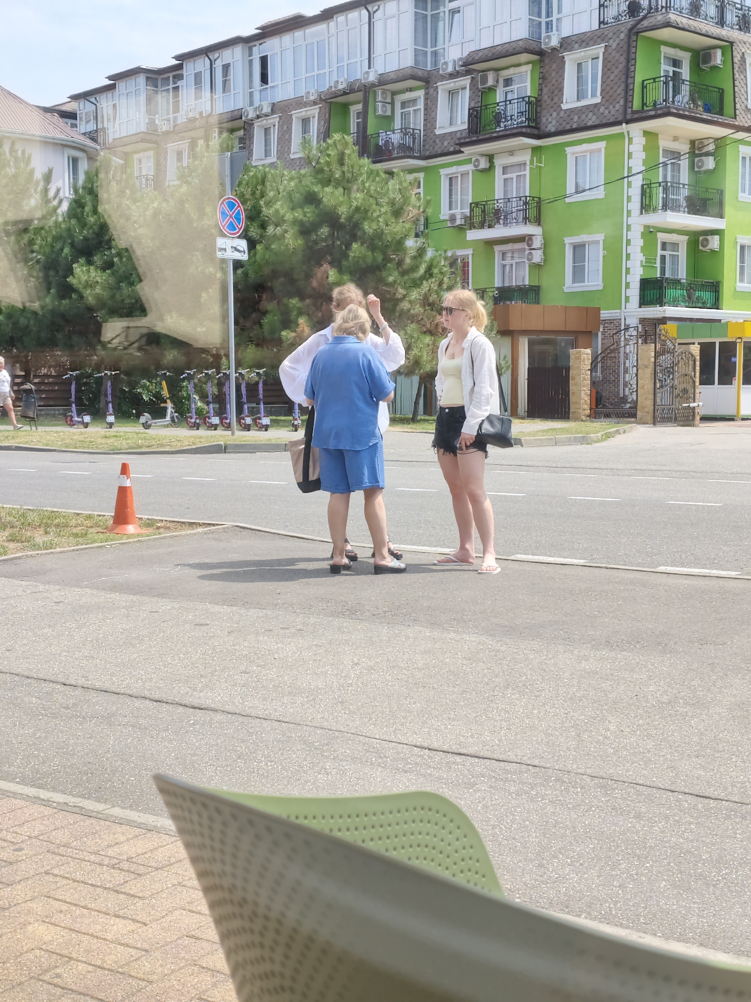 The image depicts a street scene in Sochi, Russia. In the foreground, a green plastic chair is partially visible. In the middle ground, three individuals are standing on a sidewalk near a road. One person is wearing a blue top and shorts, another a white top and dark shorts, and the third a white top and light-colored shorts. They appear to be conversing.
Behind them, a row of electric scooters is parked along a fence. The background features a brightly colored, multi-story building with green and white facades, balconies, and numerous windows. Trees and a street sign are also visible. The overall scene suggests a warm, sunny day in a developed urban area.