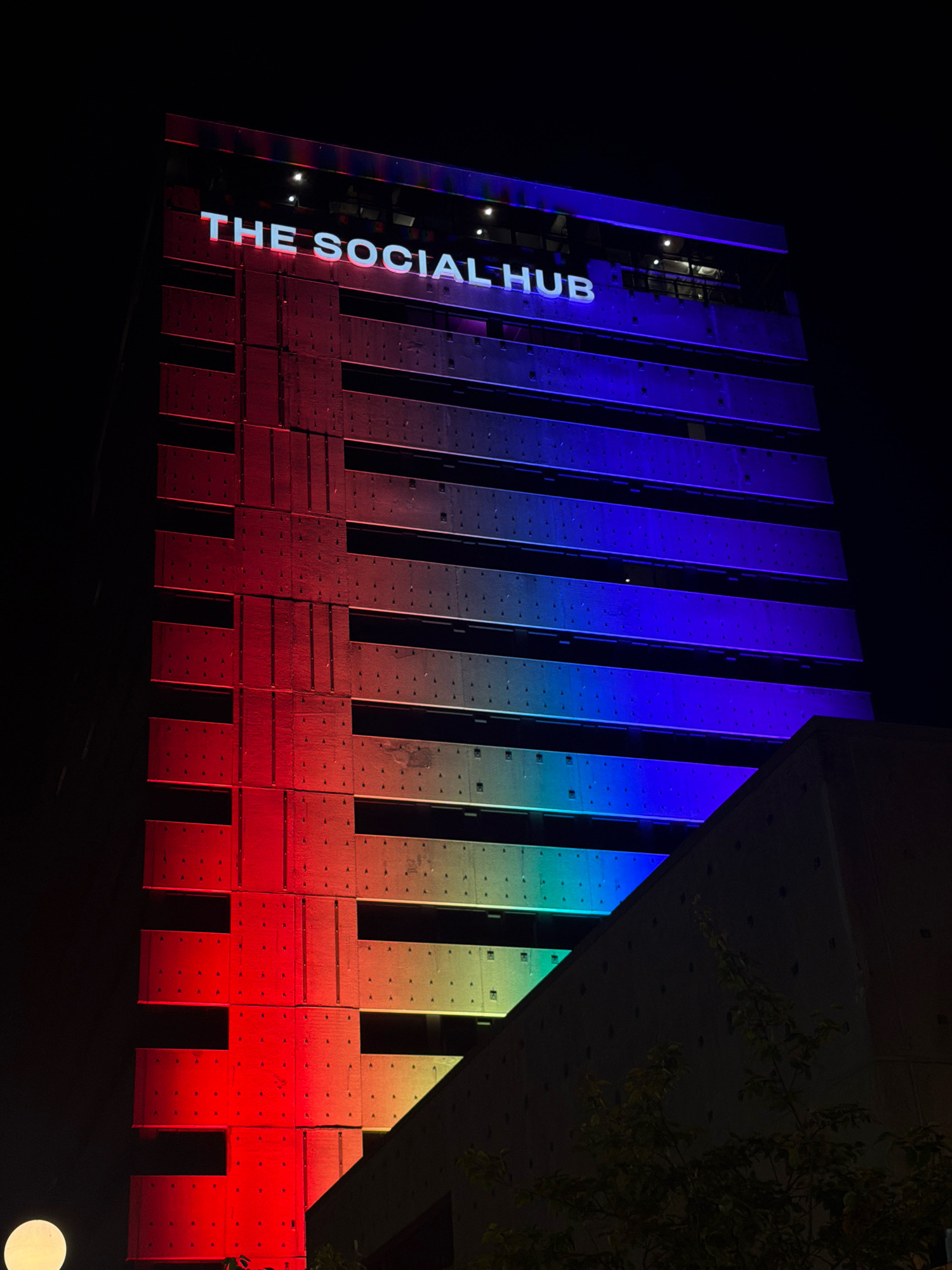 The image shows a tall building illuminated at night with a rainbow gradient of lights. The words "THE SOCIAL HUB" are displayed in white lettering on the upper facade of the building. The building's structure is visible with horizontal bands of light and small, dark circular elements on its surface, possibly windows or decorative features. In the lower left corner, a white, circular light source is visible. A portion of another building or structure, darker in color, is in the foreground on the right, with some trees and foliage in front of it. The scene appears to be an exterior view of a city building at night, with a vibrant lighting display. The building is located in Amsterdam, Netherlands.