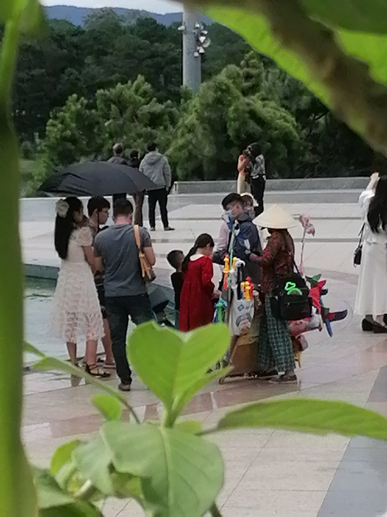 The image captures a bustling scene in a city, likely Da Lat, Vietnam, given the presence of a conical hat and the lush greenery in the background. In the foreground, a person is holding a black umbrella, partially shielding a group of individuals. A woman in a white lace dress stands to the left, while a man in a blue shirt and dark pants is positioned beside her. Further into the scene, a person wearing a conical hat appears to be selling items from a cart laden with colorful toys. The environment suggests an outdoor public space, possibly a park or a market, with paved ground and trees adorning the background. The overall impression is one of everyday life and commerce in a lively urban setting.