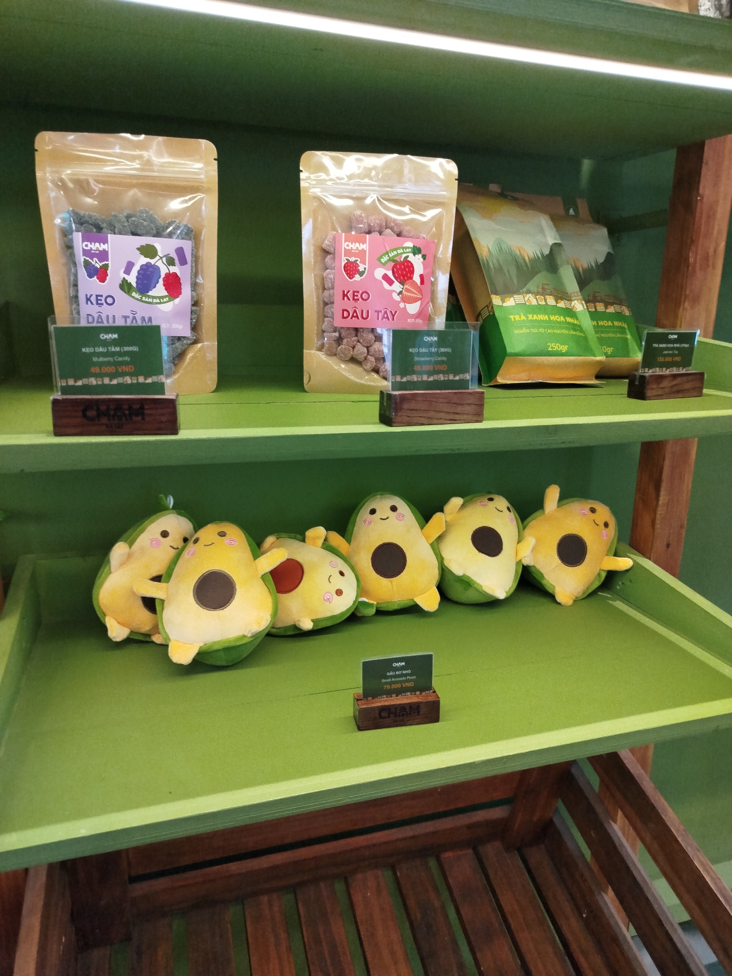 The image shows two shelves in a shop, most likely located in Da Lat, Vietnam, based on the Vietnamese writing on the product labels. The top shelf displays various products, including two clear bags of "CHAM" brand candy: "Kẹo dâu tằm" (Mulberry Candy) and "Kẹo dâu tây" (Strawberry Candy). There are also two bags of "TRÀ XANH HOA NHÀI" (Jasmine Tea). The bottom shelf features several plush avocado toys with smiling faces. Price tags are visible in front of each product, written in Vietnamese Dong (VND). The shelves are painted green and appear to be part of a wooden structure.