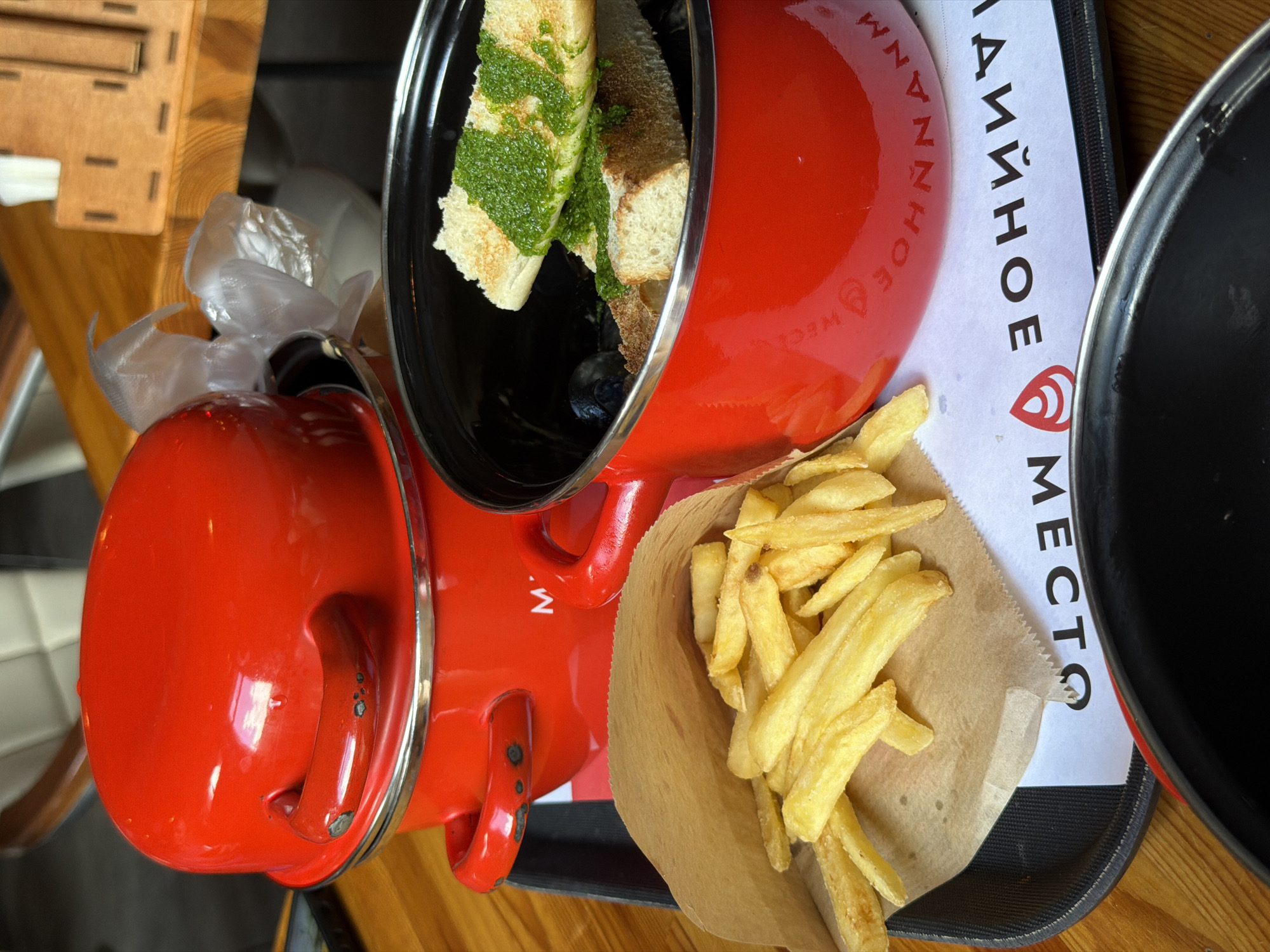 A red enamel pot with a lid is stacked on top of another identical red enamel pot. Both pots have handles. In front of the pots, a portion of a black tray is visible. On the tray, a brown paper bag filled with french fries is placed to the left. To the right of the paper bag, another red enamel pot is open, revealing a serving of what appears to be mussels topped with bread and a green sauce. The tray is on a wooden table. The background shows more wooden surfaces, a wooden container with napkins, and parts of chairs. The scene suggests a restaurant setting, possibly in Estosadok, Russia. The text "МЕСТО" is visible on a white paper underneath the tray.