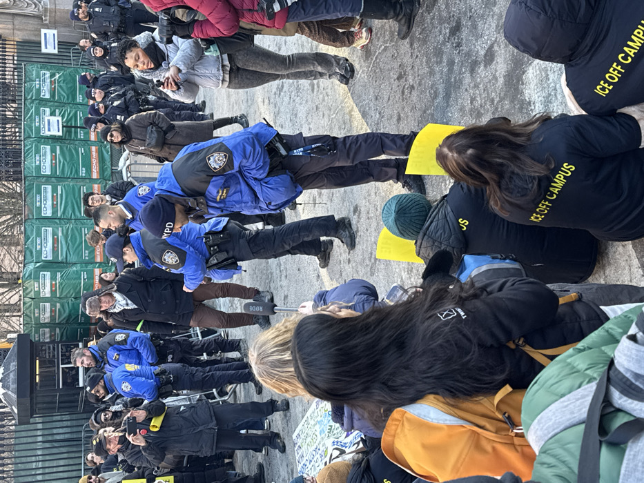 A demonstration is in progress outside a gated area with "ICE OFF CAMPUS" displayed on the backs of some participants' shirts. NYPD officers are present, standing in line, seemingly monitoring the event. Some protesters are sitting on the ground, holding signs. A microphone is visible, suggesting media coverage. The ground appears to be paved.