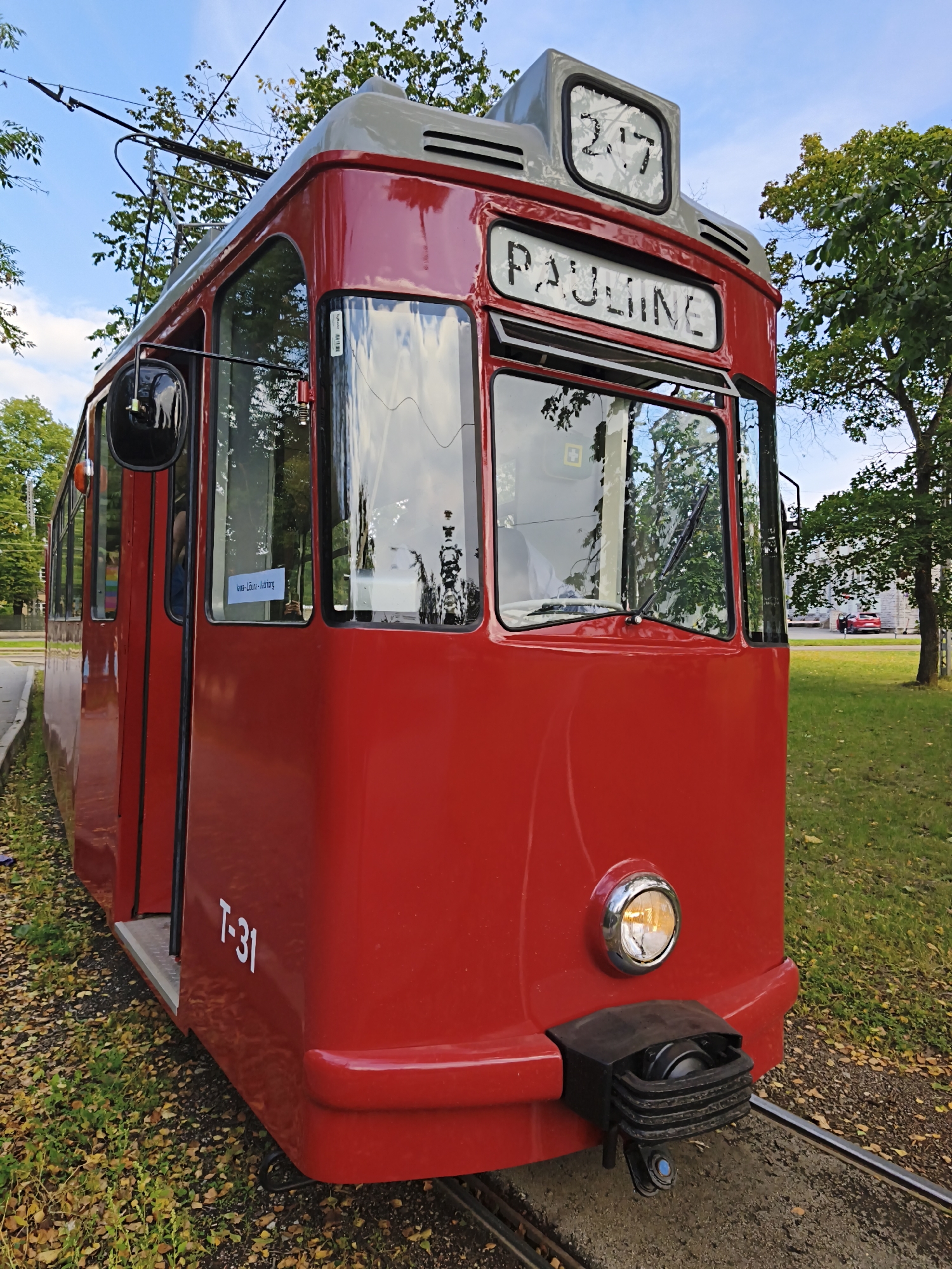 The image shows the front and side of a bright red vintage tram, labeled "PAULINE" and "2.7" on its upper front panel. The tram is positioned on railway tracks outdoors, with lush green grass and fallen autumn leaves on the ground. Trees with green foliage frame the scene, and the sky above is blue with scattered clouds. The tram has large windows, a circular headlight, and the number "T-31" is visible on its side. A small blue and white sign is affixed to one of the windows, which reads "Anna-Lüne-Astberg". In the background, a street with a red car is visible. This tram is part of the Tallinn Transport Museum (Tallinna Linnamuuseum) in Tallinn, Estonia.