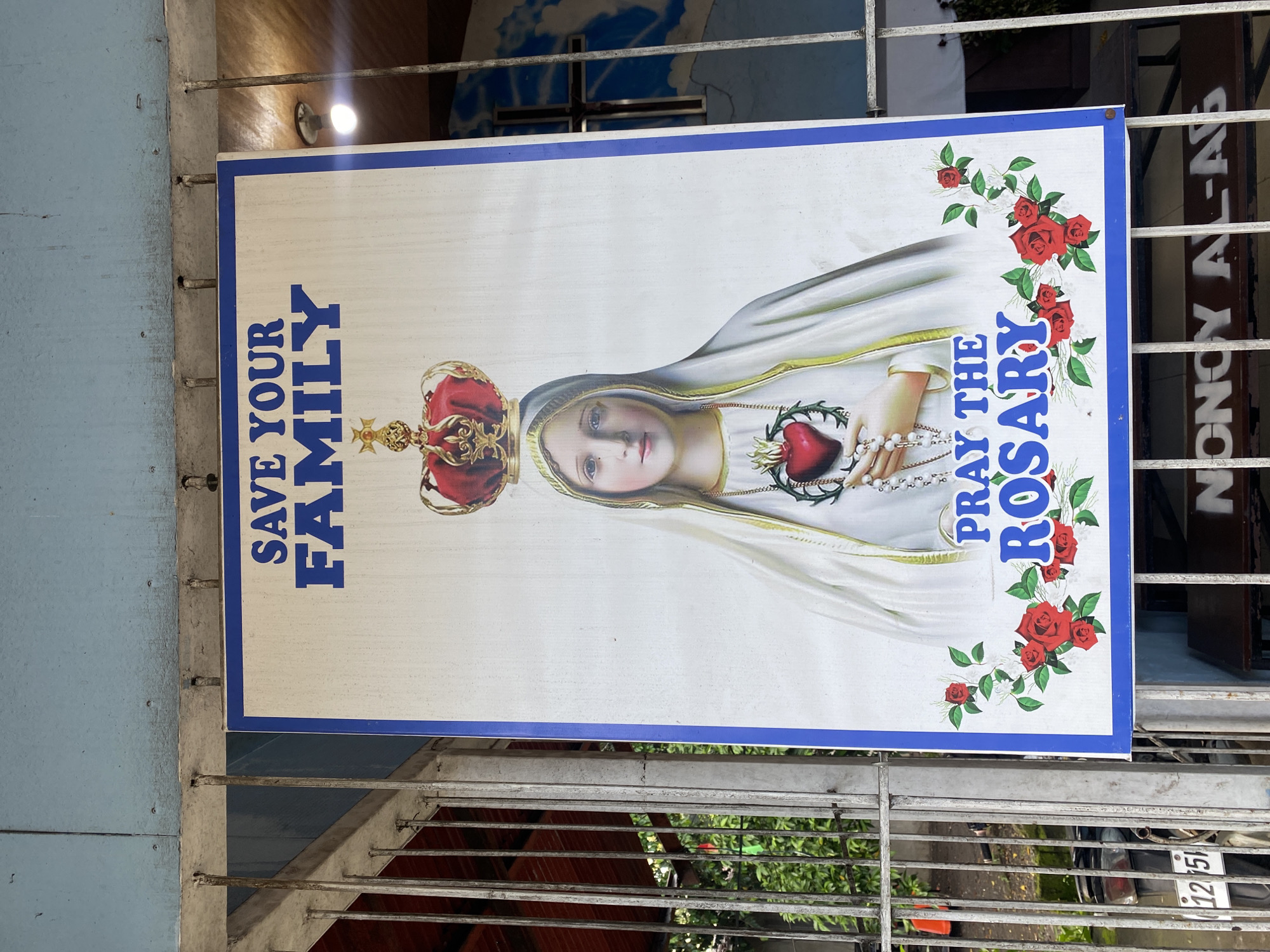A religious poster is displayed outdoors, attached to a metal fence, in what appears to be a public area. The poster features a central image of the Virgin Mary wearing a crown and a veil, holding a flaming heart with thorns, and a rosary. The text at the top reads "SAVE YOUR FAMILY" and at the bottom, "PRAY THE ROSARY." Red roses and green leaves frame the bottom portion of the poster. The poster is attached with metal fasteners to a gray, weathered metal fence. Behind the fence, a building with a light blue facade and a red-tiled roof is visible. To the right of the poster, a framed crucifix with a painted sky background can be seen through the fence. In the lower left corner of the poster, a license plate with the numbers "12751" is partially visible on a parked motorcycle. The ground beneath the fence appears to be paved. The environment suggests a tropical climate, likely in a community setting. The poster itself is slightly creased and shows signs of outdoor exposure. The overall scene conveys a message of faith and family preservation. Based on a known street art location in Davao City, Philippines, this poster is likely situated near the Roxas Avenue Boulevard.