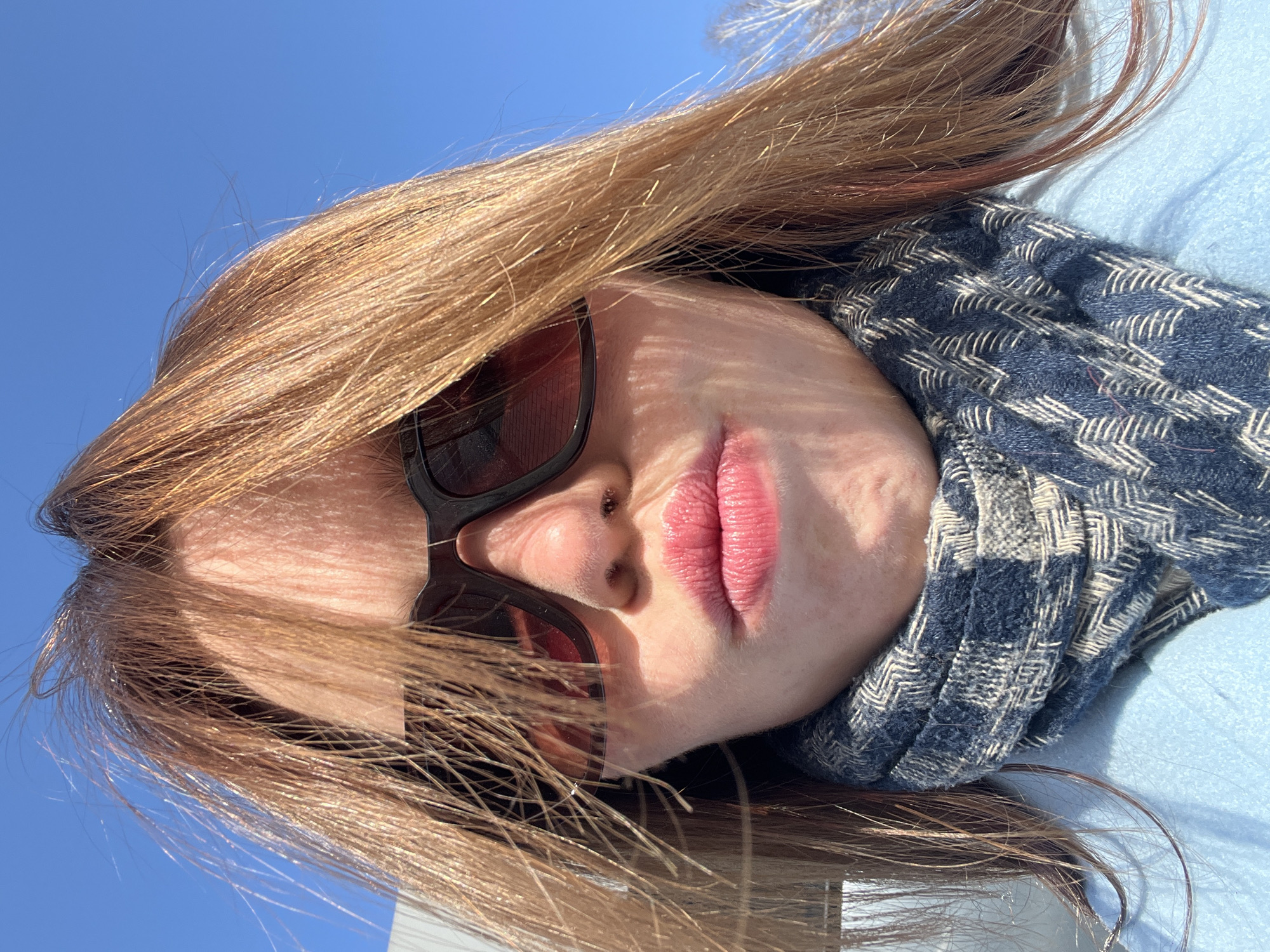 A close-up outdoor portrait captures an individual's head and upper torso from a slightly upward angle, against a clear blue sky. The person has light brown, windswept hair, illuminated by sunlight that creates a golden sheen. Dark-framed sunglasses with dark, reddish-tinted lenses obscure the eyes and cast distinct shadows on the upper cheeks. A small piercing is visible on the left nostril. The lips are pursed in a 'kissing' expression and are colored with a pink hue. A thick, textured scarf featuring a geometric pattern of blue and cream-colored lines is wrapped around the neck, worn over a light blue coat. In the background, a light-colored structure is partially visible on the left, and bare tree branches are discernible against the sky on the far right. The image depicts a scene in the city of Chaikovsky, Russia.