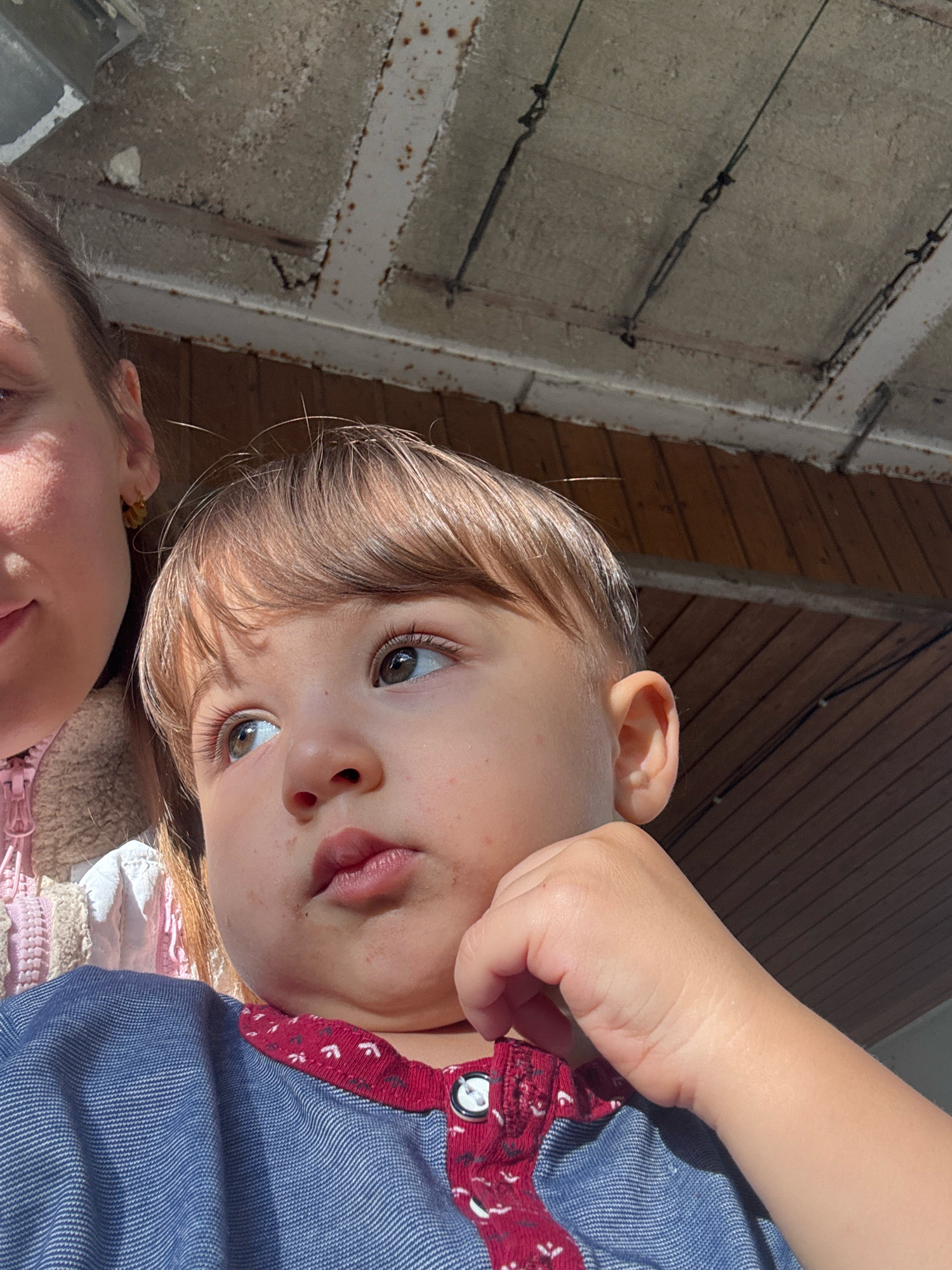 An outdoor, close-up, eye-level, medium shot captures a young child from the chest up, looking to the right, with a partial view of an adult on the left. The child has light brown hair, green eyes, and is resting their right hand on their chin. They are wearing a blue shirt with a red and white patterned collar and buttons. The adult, partially visible on the left, appears to be a woman with brown hair, wearing a pink jacket. The background consists of a wooden ceiling structure with visible beams and what appears to be a corrugated metal roof or overhang. The lighting suggests it is daytime, with sunlight casting shadows. The environment appears to be an outdoor sheltered area, possibly a porch or a covered patio, in Überherrn, Germany.