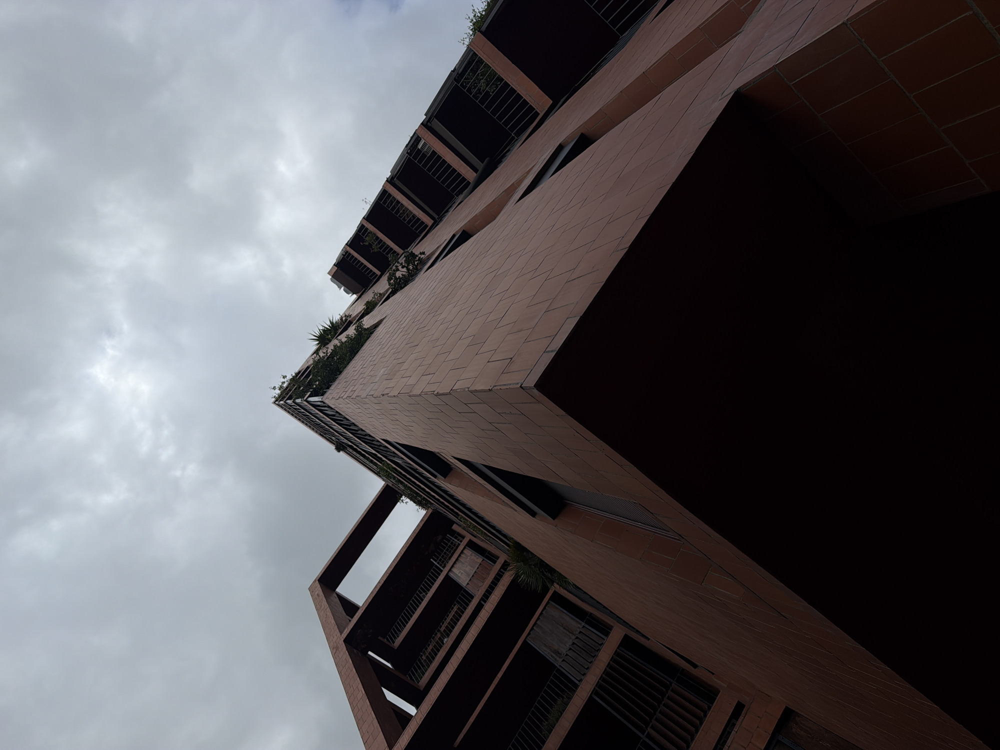 An extreme low-angle, wide shot captures the exterior of a modern terracotta-clad building against a cloudy sky. The building, situated in the Poble Sec neighborhood of Barcelona, Spain, features multiple balconies with dark metal railings and some sparse greenery. The architecture is characterized by sharp angles and cantilevered sections, creating an imposing and slightly dramatic visual. The sky is overcast and gray, contributing to a muted and somewhat somber atmosphere. No people are visible in the frame.