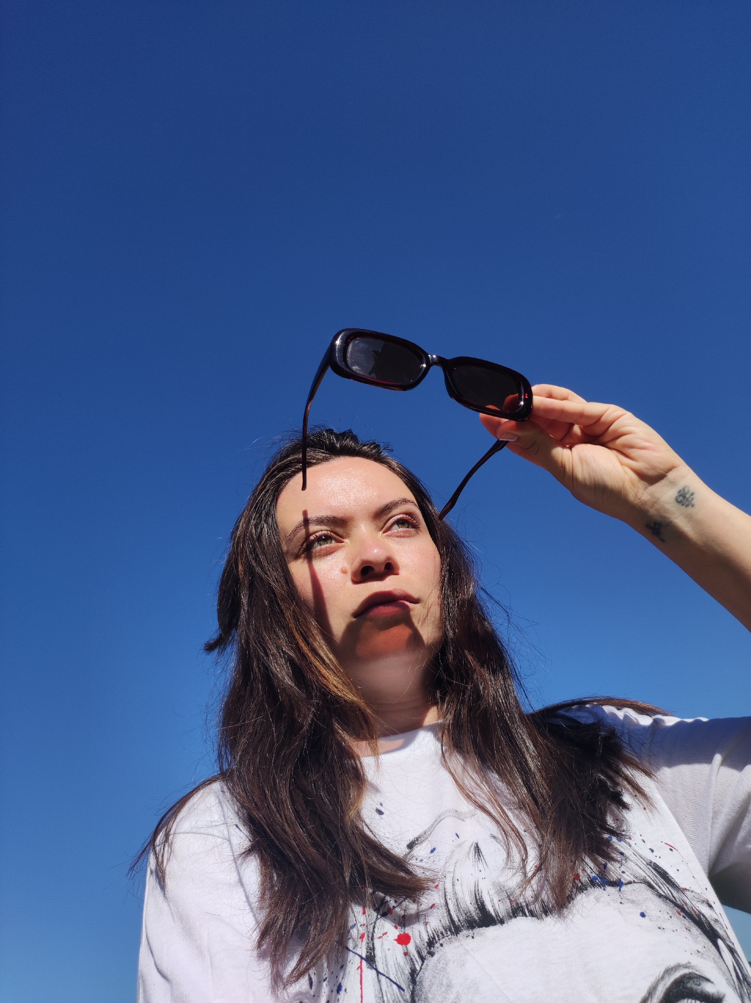 A woman holds up a pair of sunglasses. The background is clear blue sky. The woman is wearing a white t-shirt with a black and white graphic print and her hair is long and brown. The tattoo on her right wrist is visible.
There is no information in the picture to indicate it was taken in the city of Фоки, Russia.