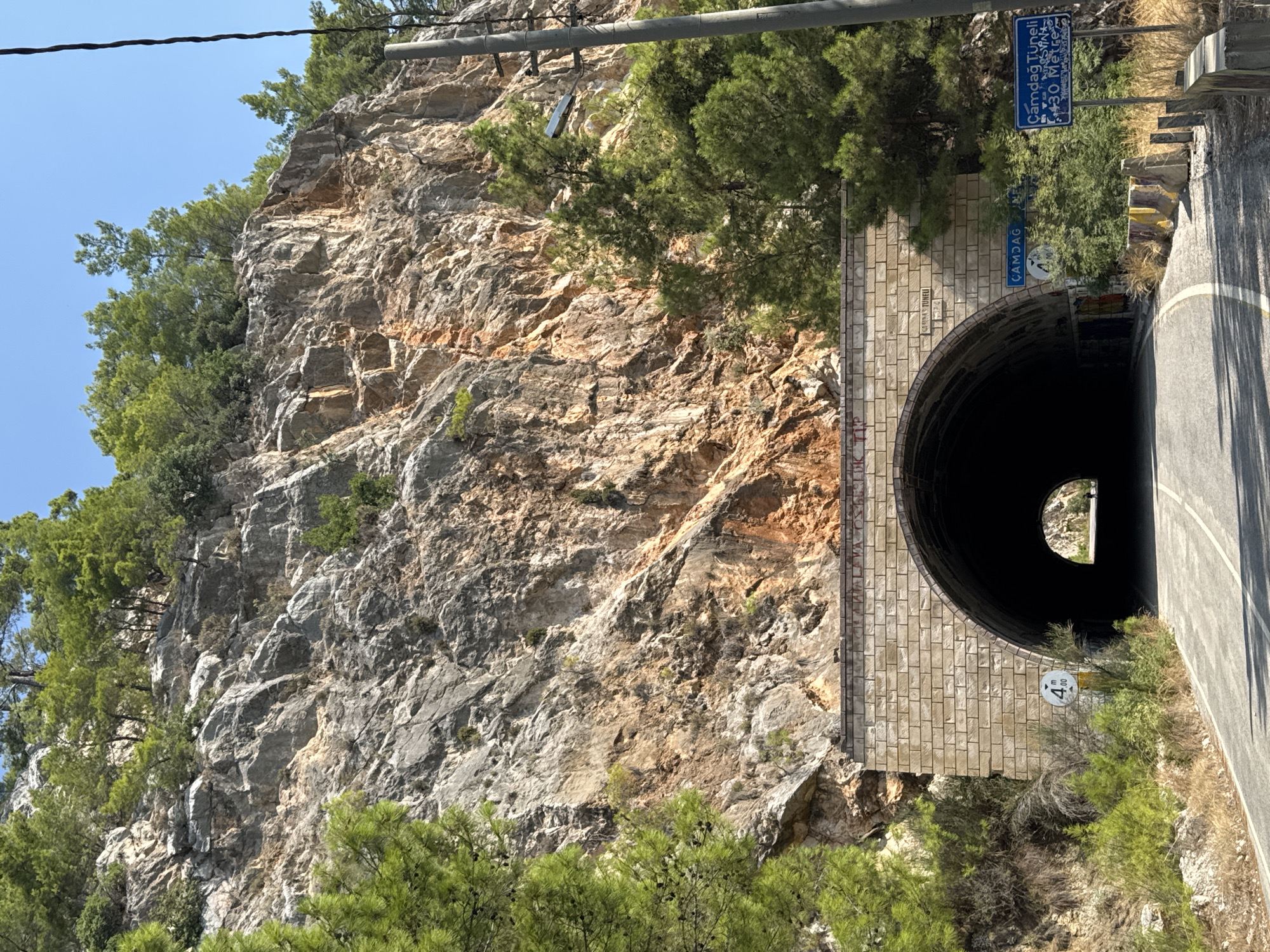 The image shows the entrance to the Çamdağ Tunnel on a sunny day. The tunnel is carved into a rocky mountainside covered with trees. The entrance is made of light-colored stone blocks. A sign to the right of the tunnel entrance reads "Çamdağ Tüneli 430 Metre," indicating the tunnel's name and length. A road leads into the dark tunnel, with a sign showing a height limit of 4.00 meters on the left side of the road. The surrounding environment is dry and rocky, with sparse vegetation. The sky is clear and blue. No people are visible in the image.