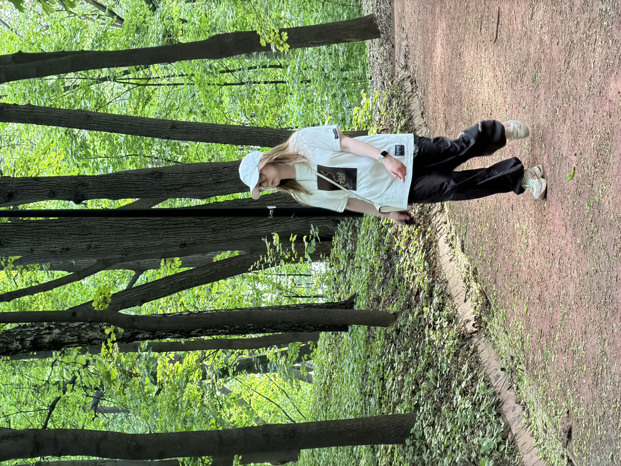 The image shows a person walking on a path in a park or wooded area, likely in Moscow, Russia, given the presence of deciduous trees and the general appearance of a managed green space. The individual is wearing a white baseball cap, a light-colored t-shirt with a dark graphic, dark trousers, and light-colored sneakers. They appear to be holding a phone or small device in their hand. The path is unpaved, likely dirt or gravel, bordered by dense foliage and trees. The lighting suggests it is daytime, with dappled sunlight filtering through the trees.