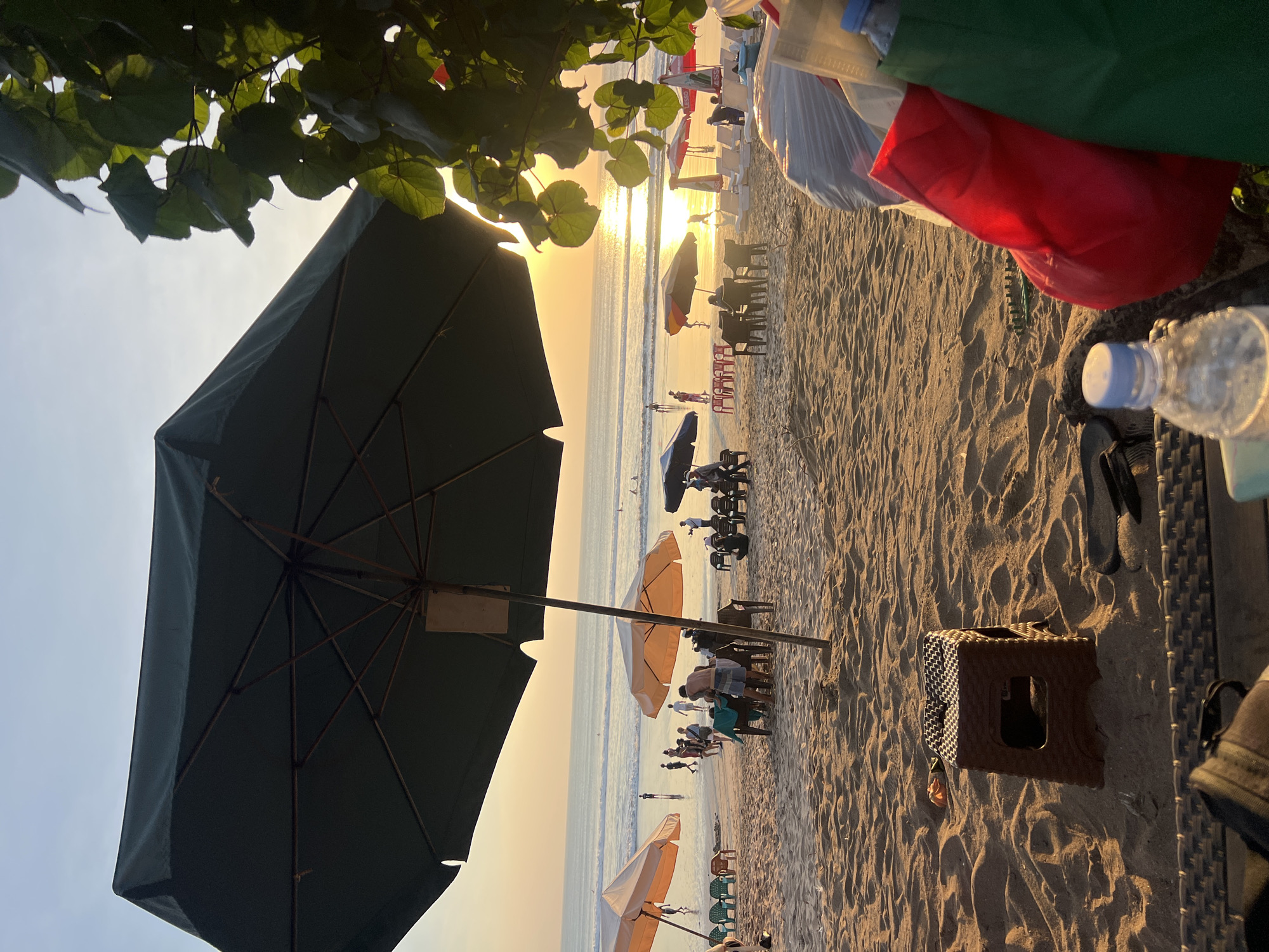 In Kuta, Indonesia, this image captures a beach scene during sunset. The foreground features a large, dark green beach umbrella, casting a shadow over the sandy ground. To the right, a pile of bags, including a red one and a green one, are visible, along with a plastic water bottle. Further back, a brown plastic stool sits on the sand.
The midground of the image reveals more beach umbrellas, some open and casting shade, with beach chairs scattered around them. Several people are walking along the shore and gathered at tables under the umbrellas. The ocean stretches out to the horizon, with gentle waves and a few distant figures swimming or surfing.
The sky is a warm gradient of orange and yellow, indicating the sun is setting. The overall atmosphere is relaxed and depicts a typical evening on a popular tourist beach.