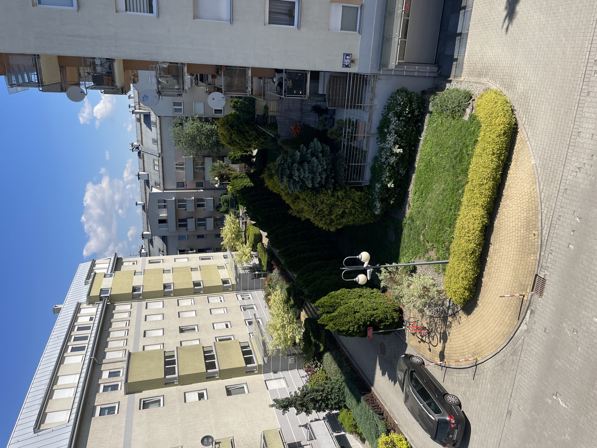 This is an elevated daytime view of a residential courtyard area within an urban setting. The location is in Unknown, Unknown. Several multi-story apartment buildings border a well-maintained courtyard. The courtyard features a variety of landscaping elements, including hedges, lawn areas, trees, and flowering plants. A curved pathway made of yellow paving stones runs through a portion of the courtyard. A parked black car is visible on the pavement, along with a red bicycle leaning against a lamp post. The sky is blue with scattered white clouds. Shadows indicate it is daytime, and the sun is positioned to the upper right of the frame.