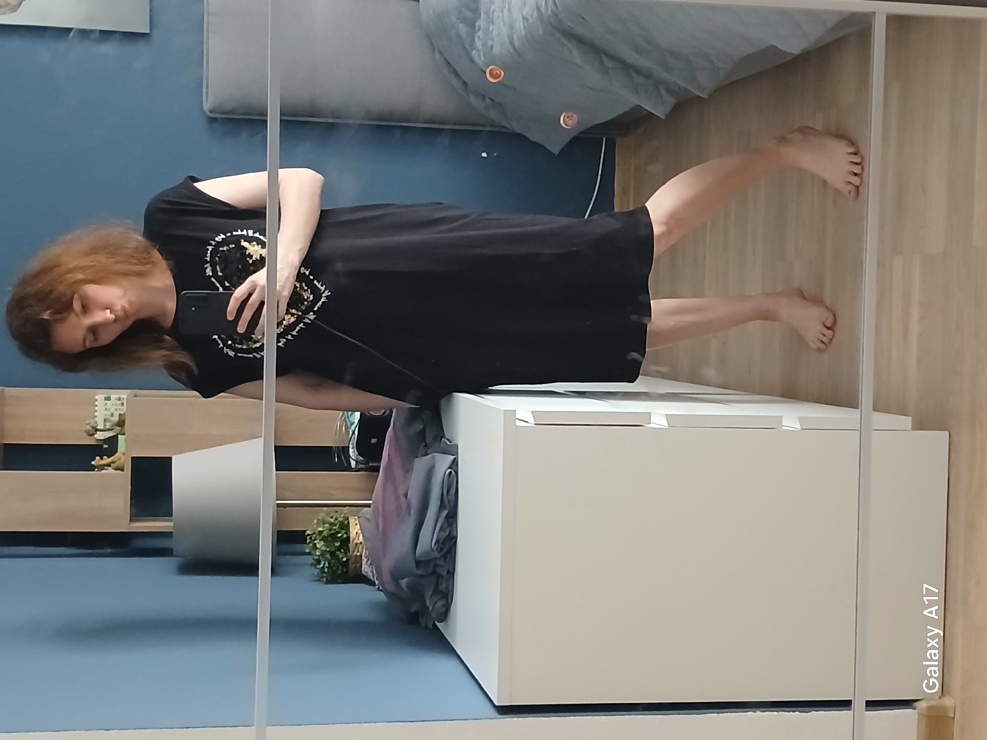 A woman with reddish-brown hair is standing in front of a mirror, holding a smartphone and taking a picture of herself. She is wearing a black dress with a decorative pattern. The mirror reflects the room, showing a bed with grey bedding and a blue wall. To the left of the woman, there is a shelving unit with decorative items. A white dresser is also visible, with some clothes draped over the side. The flooring appears to be light-colored wood. The location may be a private residence in Yekaterinburg, Russia, given the domestic setting, but there is no specific visual evidence to confirm this.