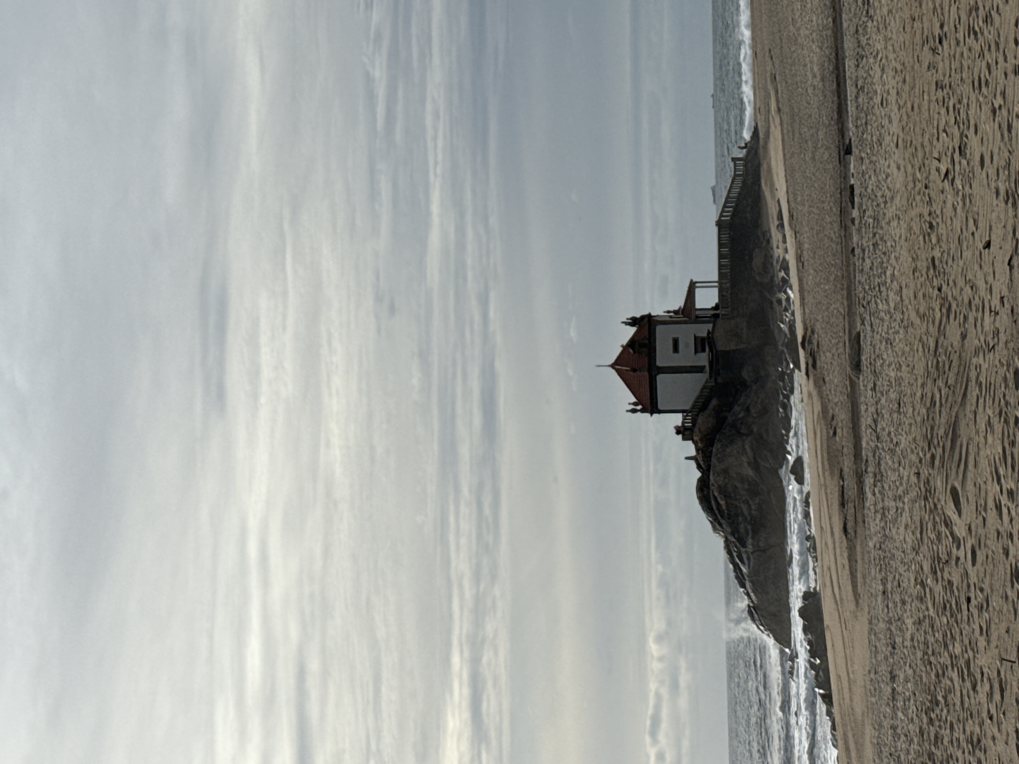 The image captures the Capela do Senhor da Pedra (Chapel of the Lord of the Rock) in Vila Nova de Gaia, Portugal, at the Praia de Miramar. The chapel sits on a rocky outcrop along the shoreline, a small, white building with a red-tiled roof. The Atlantic Ocean is visible, with waves washing against the rocks beneath the chapel. A sandy beach stretches along the right side of the frame. The sky is overcast, with light, striated clouds spanning the upper half of the image. No people are visibly present.