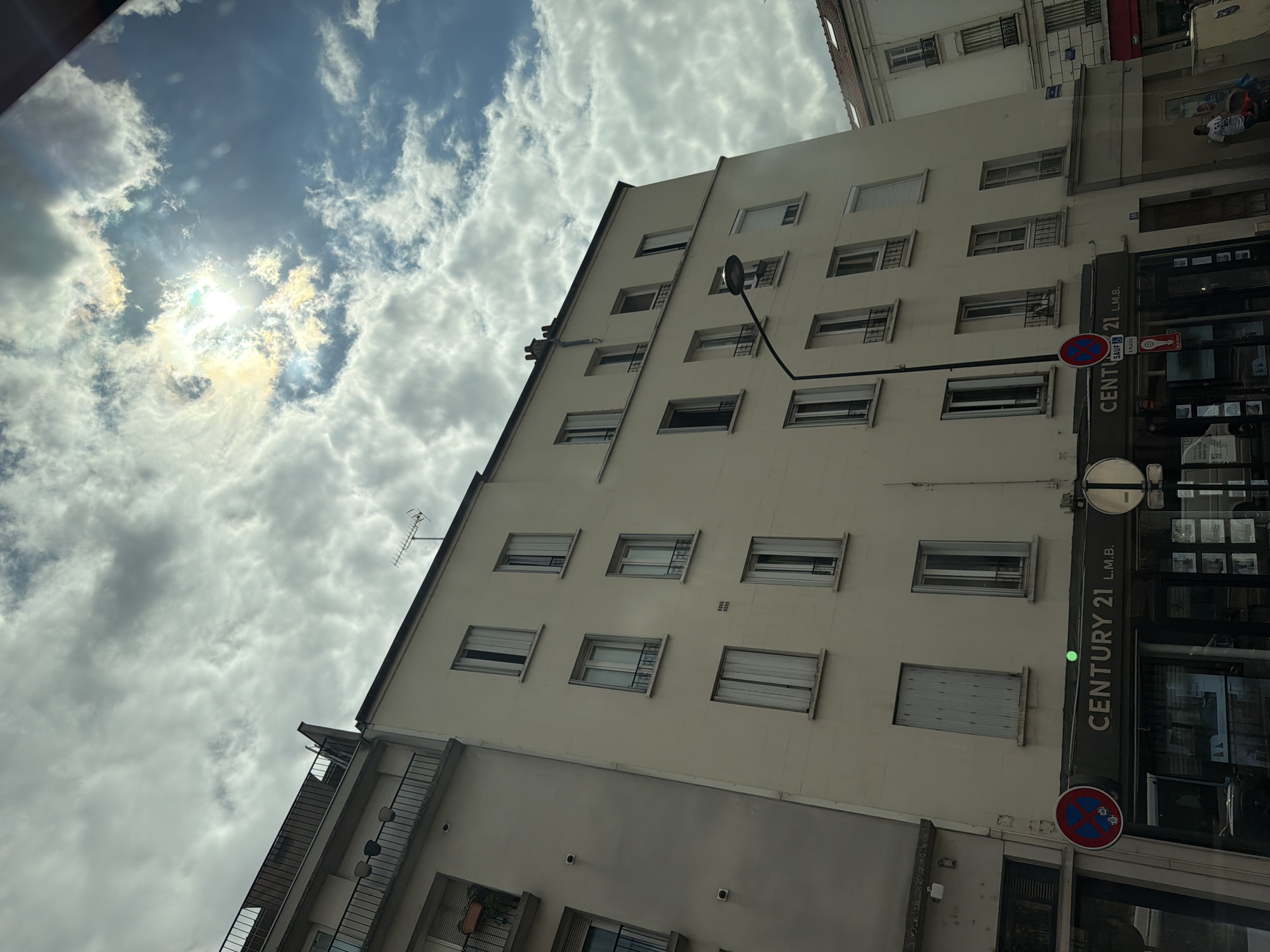 This media file depicts a view of buildings and the sky, taken from an upward-angled perspective. The primary subjects are the facades of residential or commercial buildings, characterized by multiple windows and architectural details such as balconies and external lighting fixtures. On the right side of the image, a vertical sign for "CENTURY 21 L.M.B." is prominently displayed, indicating a real estate agency. Below this, a circular "no parking" sign is visible.
The sky is filled with scattered clouds, suggesting a daytime scene with variable weather conditions. Sunlight breaks through in some areas, creating bright spots and illuminating the clouds. The overall impression is of an urban environment.
While the image is primarily focused on the architecture and sky, a person can be seen through a window in the background, suggesting indoor activity. The scene appears to be set in Alfortville, France, as indicated by the provided location context. The lighting suggests it is daytime, possibly late afternoon or early morning given the angle of the sun. No specific emotions are conveyed, and the activity is primarily static observation of the urban landscape.