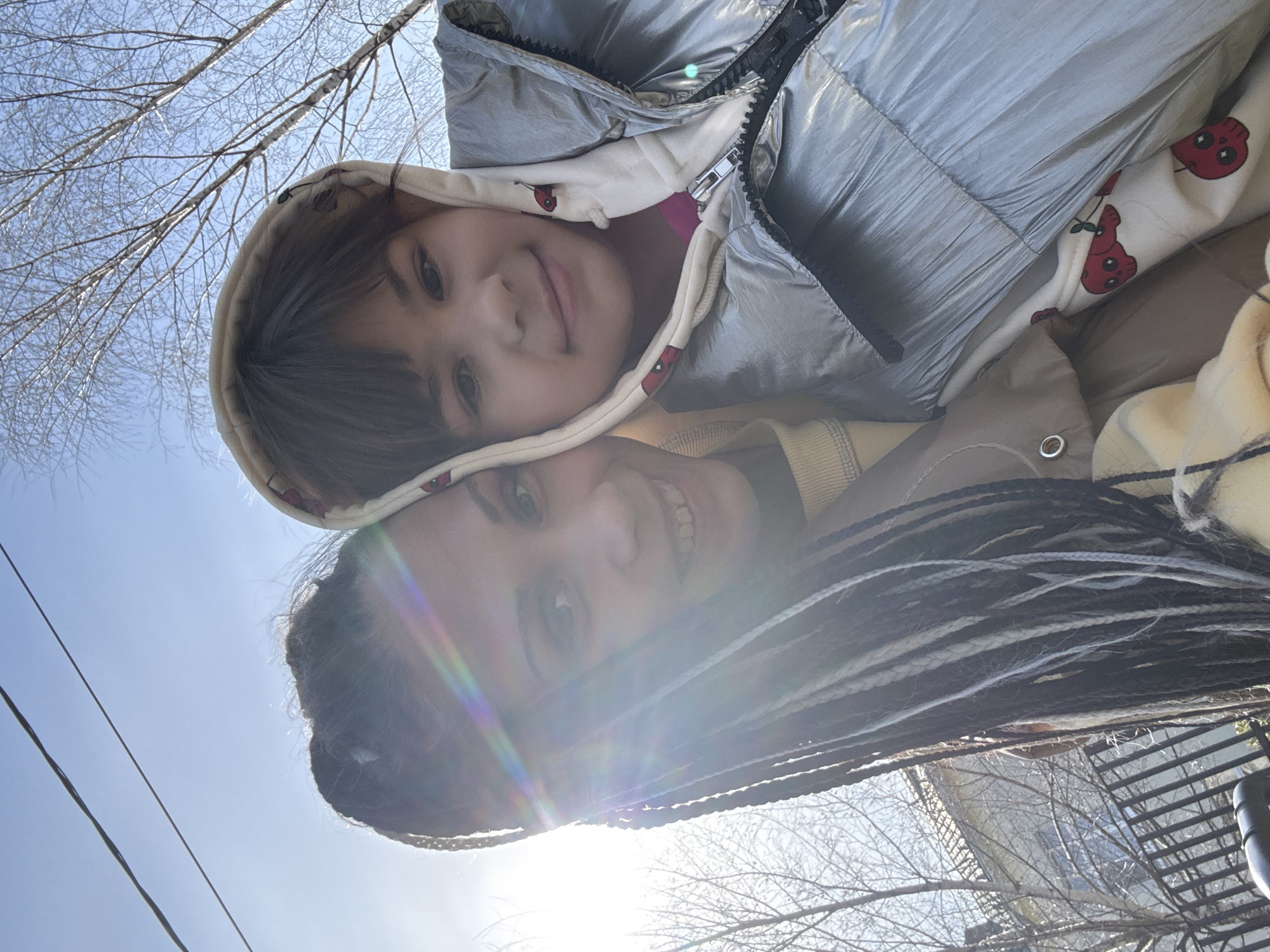 A low-angle selfie depicts a woman and a child outside. The child, wearing a hooded jacket with cartoon cherry graphics and a shiny silver puffer vest, is positioned above the woman. The woman has dark hair styled in long braids with white streaks and is smiling broadly. The background includes bare tree branches against a bright blue sky with a strong solar flare. The location is likely Rusanovo, Russia, as specified in the prompt. Additional details visible include clothing fasteners on the child's jacket, overhead power lines, and glimpses of nearby buildings and fencing.