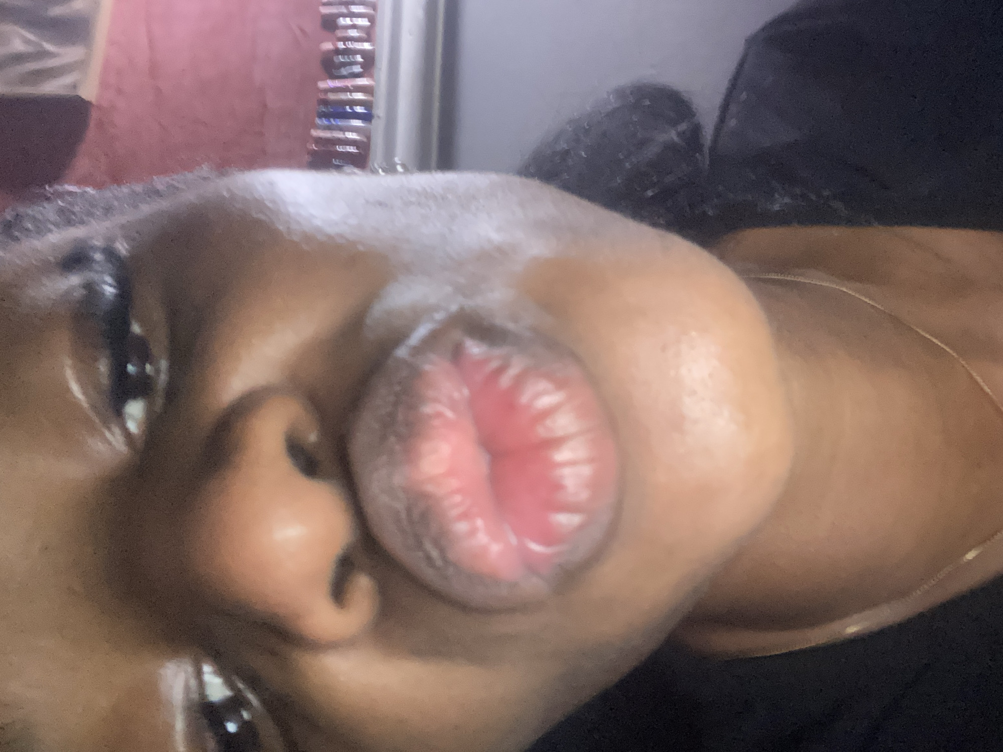 The image is a close-up, selfie-style photograph of a person's face, with a particular focus on their lips which are pursed. The person appears to be wearing makeup, including mascara on their eyelashes and a pinkish lipstick. They are also wearing a simple gold chain necklace. The background is out of focus and suggests an indoor setting, possibly a room with a window covered by blinds or curtains, and a shelf or surface with items on it. There is no indication of the location being the city of Jos, Nigeria.