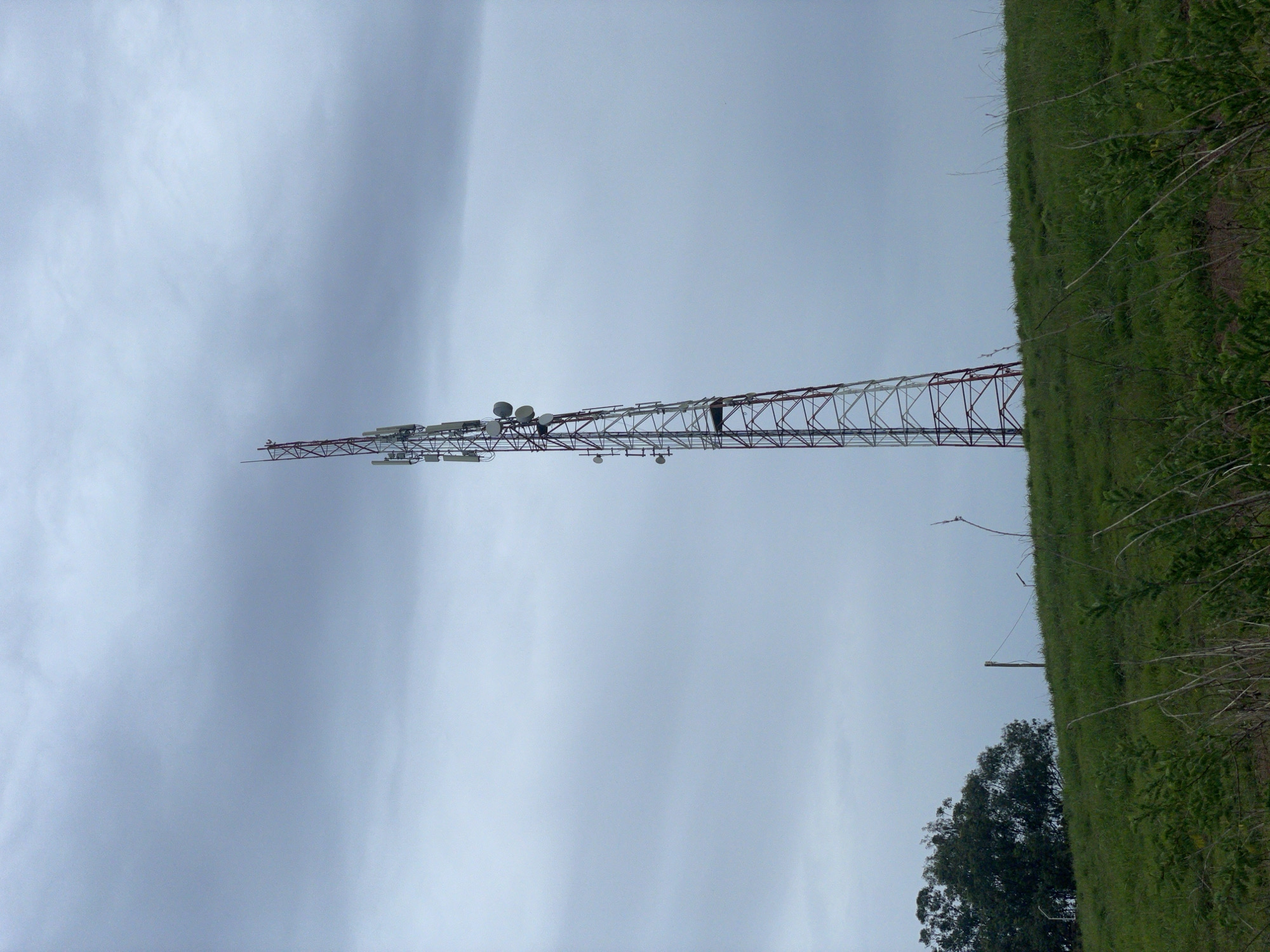 The image shows a tall communication tower made of red and white lattice metal. The tower is adorned with several antennas and other equipment at different levels. A person is visible on the tower, appearing to be working on it. The tower stands in a grassy, hilly landscape. The sky is overcast with thick, grey clouds, suggesting a cloudy or possibly rainy day. The overall scene conveys an industrial or telecommunications setting in a rural or semi-rural environment. The grass on the hill is green and appears lush. There are some trees and branches visible in the foreground and background, on the right side of the image. The image is oriented vertically, with the tower appearing to stretch upwards from the bottom of the frame to the top. There is no visible text in the image. The location is identified as Durimi, Nigeria.