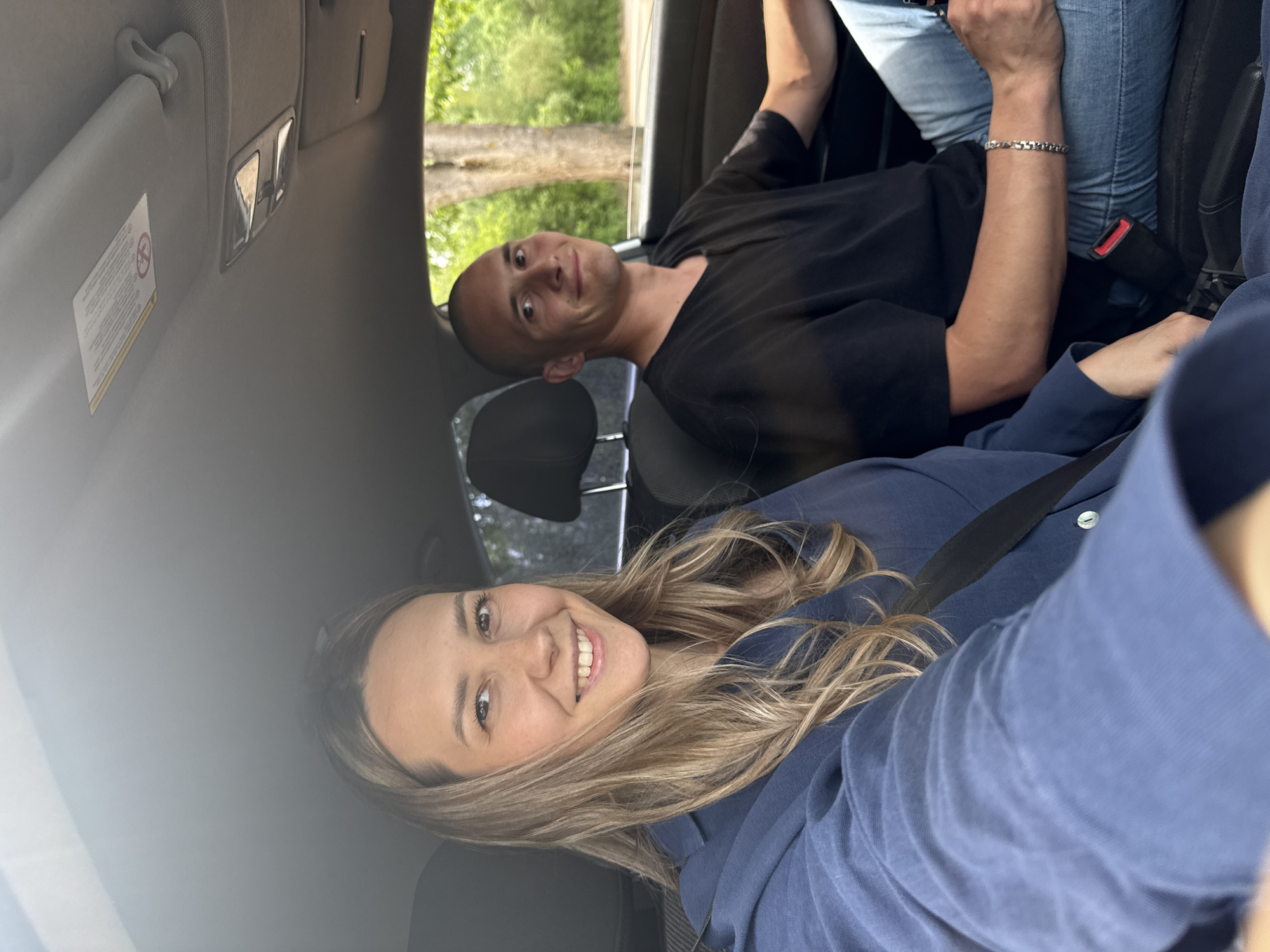 Two individuals are seated in a car. A woman with blonde hair is in the foreground, wearing a blue shirt and a seatbelt. She is looking at the camera and smiling. Behind her, a man with a shaved head is wearing a black t-shirt and is also looking at the camera with a slight smile. The car is moving, as suggested by the blurred view of trees and a body of water visible through the window. The interior of the car is visible, including the dashboard, the sun visor, and the car seats. The location is Chaikovsky, Russia.
