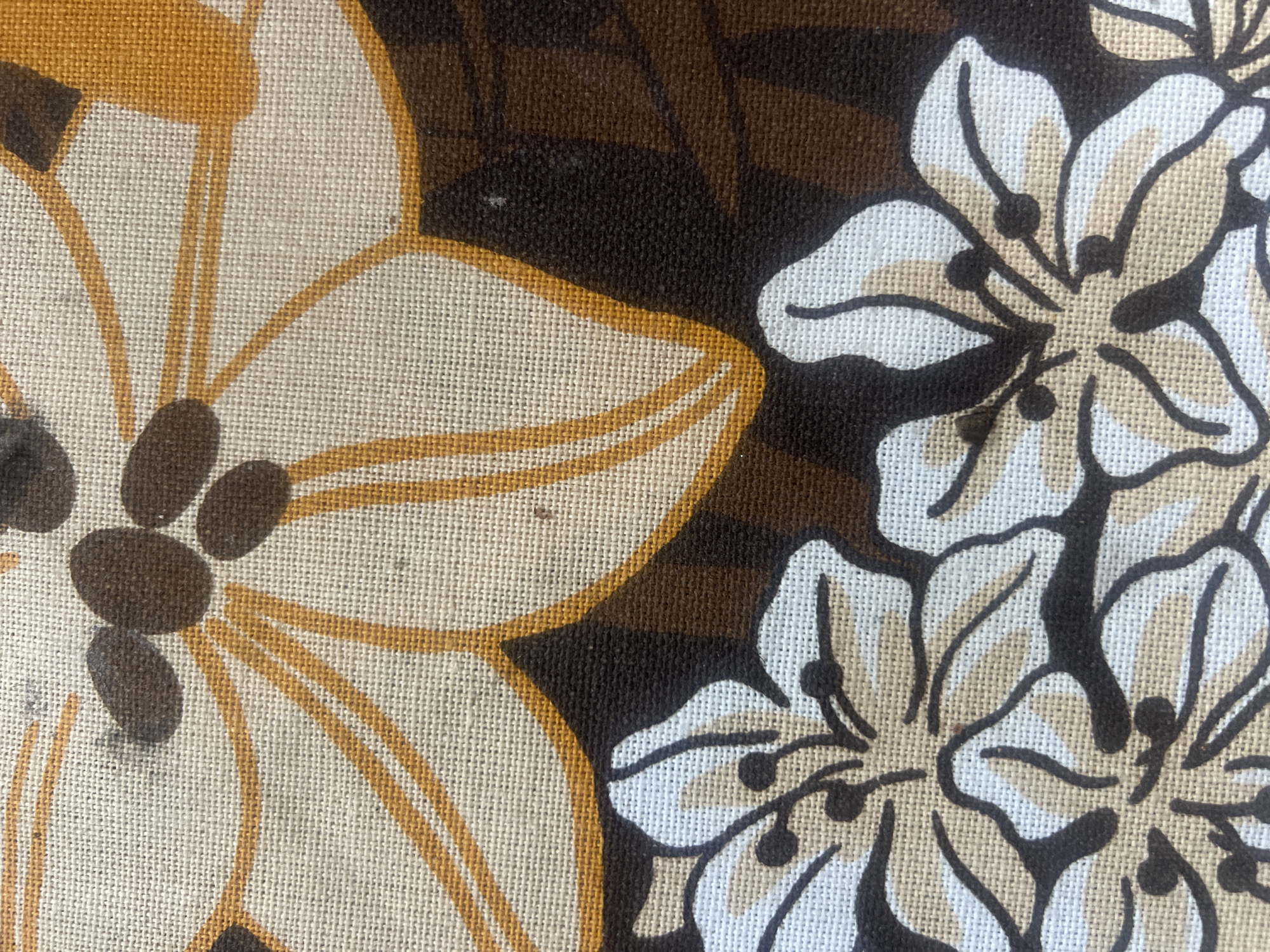The image displays a close-up, high-resolution view of a printed textile, situated in the city of Casablanca, Morocco. The fabric exhibits a woven texture, characteristic of a plain weave, likely linen or a similar natural fiber, showing discernible individual threads. The pattern consists of stylized floral motifs arranged in two distinct sections.
The upper section features large, partially visible flowers against a light beige background, which is the natural color of the fabric. These flowers are outlined in an ochre-orange hue, and their centers contain multiple dark brown, oval-shaped elements. The petals themselves are a creamy off-white or light beige.
The lower section presents smaller, more intricately detailed flowers set against a solid dark brown background. These flowers have petals rendered in both white and light beige, outlined in dark brown. Their centers feature small, dark brown circular dots, possibly representing stamens or pistils.
Minor imperfections, such as small dark specks and slight discoloration, are visible on the lighter parts of the fabric, indicating some wear or environmental dust. No people, specific actions, or direct interactions are depicted. The environment is limited to the textile surface itself.