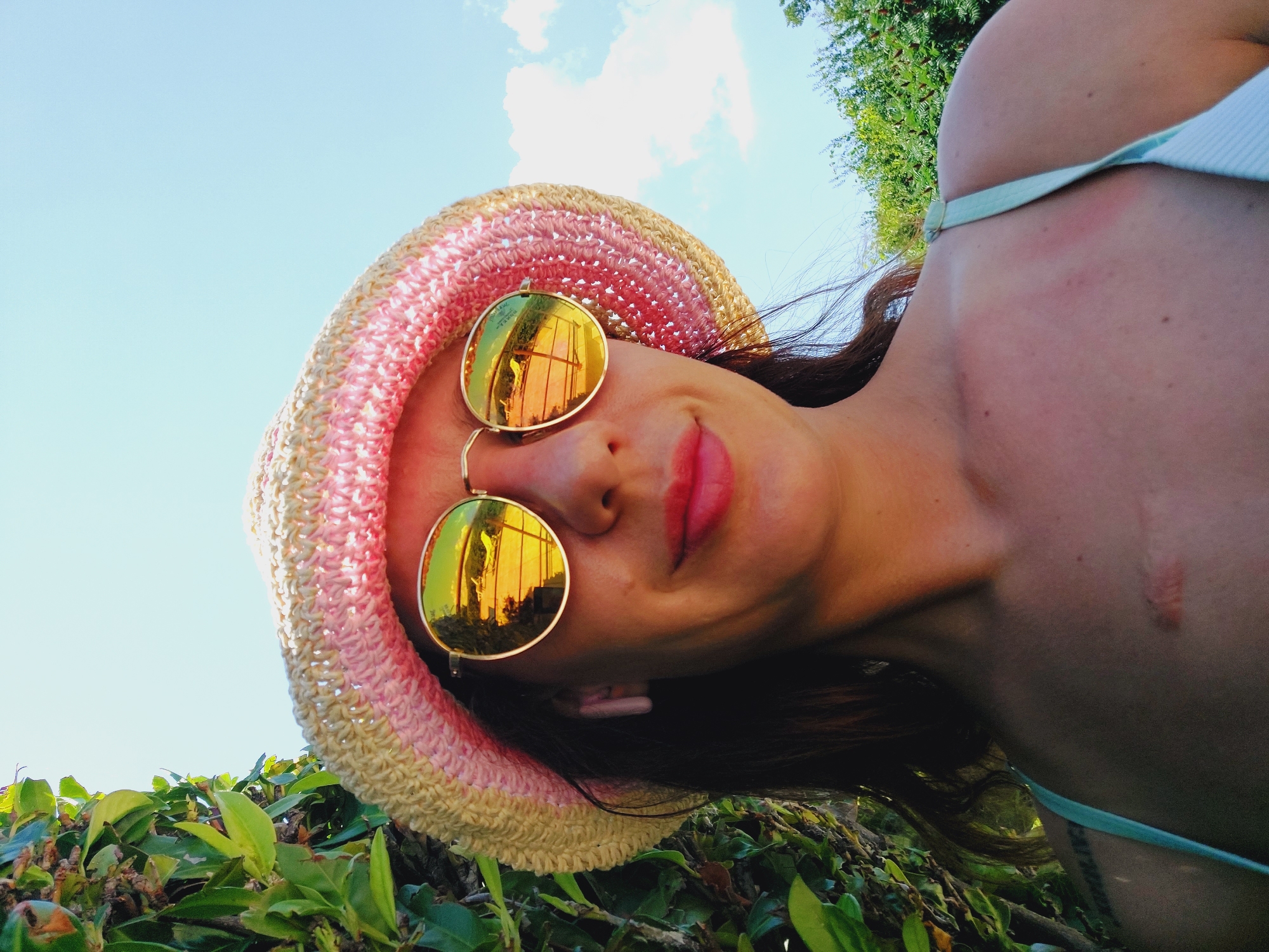 A close-up, low-angle, outdoor, daytime selfie features a woman wearing a straw hat with pink trim and reflective sunglasses. The sunglasses' lenses reflect a scene with lounge chairs and a pool. She has long, dark hair and is wearing a light blue camisole. Her chest shows a faint scar. Green foliage is visible to the left and trees are in the background to the right. The sky is clear blue with some clouds. The location is identified as Sozopol, Bulgaria.