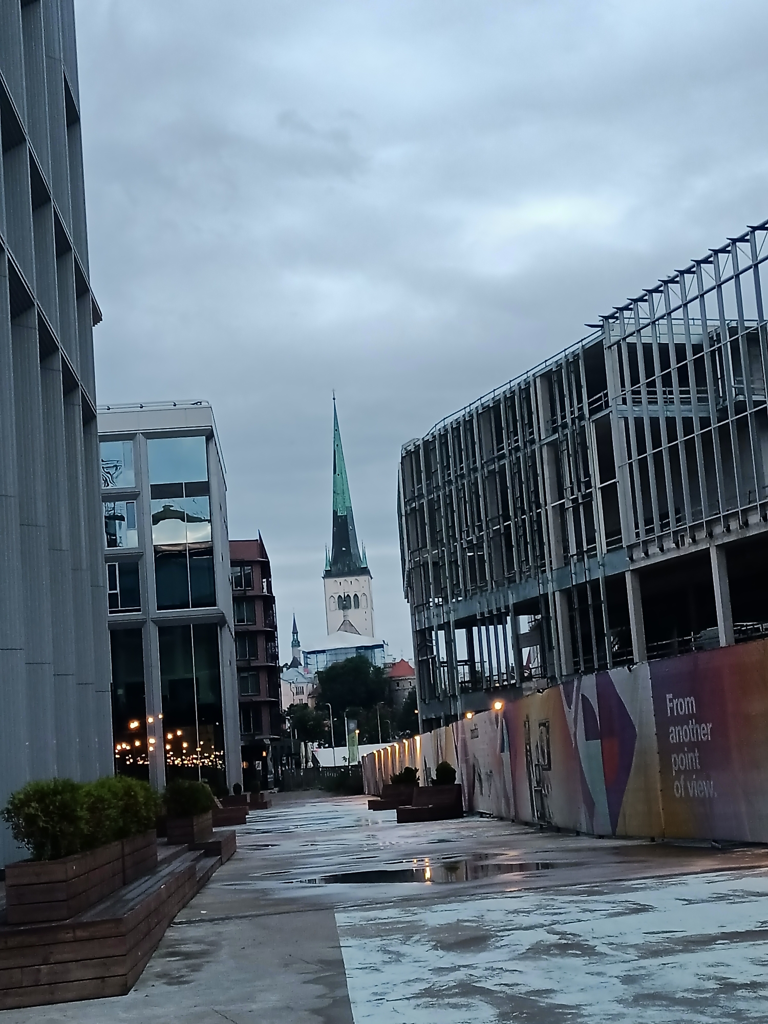 The image depicts an urban scene in Tallinn, Estonia, characterized by overcast skies and a damp, reflective ground surface suggesting recent rain. In the foreground, a modern building with a textured grey facade occupies the left side of the frame. Adjacent to it, a contemporary building with large glass windows is visible. A pathway with wooden planters containing small green bushes runs along the left side of the scene.
Further down the path, construction is evident. A partially built structure with exposed steel beams and concrete floors is on the right, enclosed by a construction barrier featuring colorful graphics and text that reads "From another point of view." In the distance, a prominent medieval-style church tower with a green spire stands tall, flanked by other buildings. The overall atmosphere is one of a city blending historical architecture with modern development, under a subdued, cloudy sky. No people are visible in the frame.