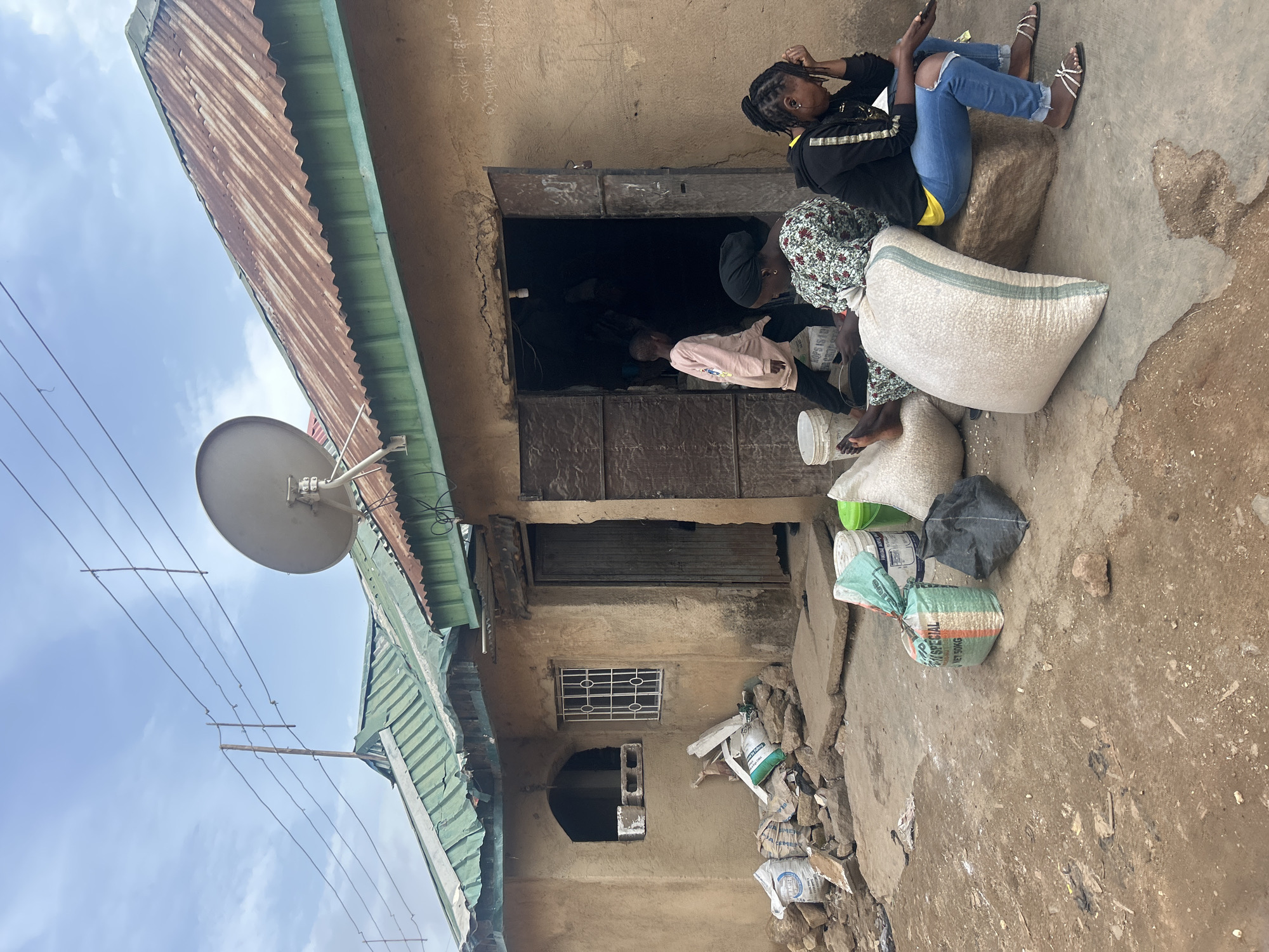 In Jos, Nigeria, on a cloudy day, an outdoor scene unfolds in front of a rustic, mud-walled building with a corrugated metal roof. The building has a large, open doorway, suggesting it might be a shop or dwelling. A satellite dish is mounted on the roof.
Several large sacks of what appear to be grain or agricultural produce are scattered around the entrance, some stacked neatly and others lying on the ground. There are also various buckets and building materials like bricks visible near the doorway. The ground is a mix of dirt and rough concrete.
Three people are present. Two women are seated on large sacks near the doorway. One woman, wearing a black headscarf and a floral patterned top, is looking downwards. The other woman, in a black hoodie and ripped blue jeans, is sitting on a large rock or sack, holding her phone and appearing thoughtful or concerned. A young child, wearing a light pink hooded jacket, is walking away from the doorway into the open area. The overall impression is one of everyday life in a semi-urban or rural setting in Jos.