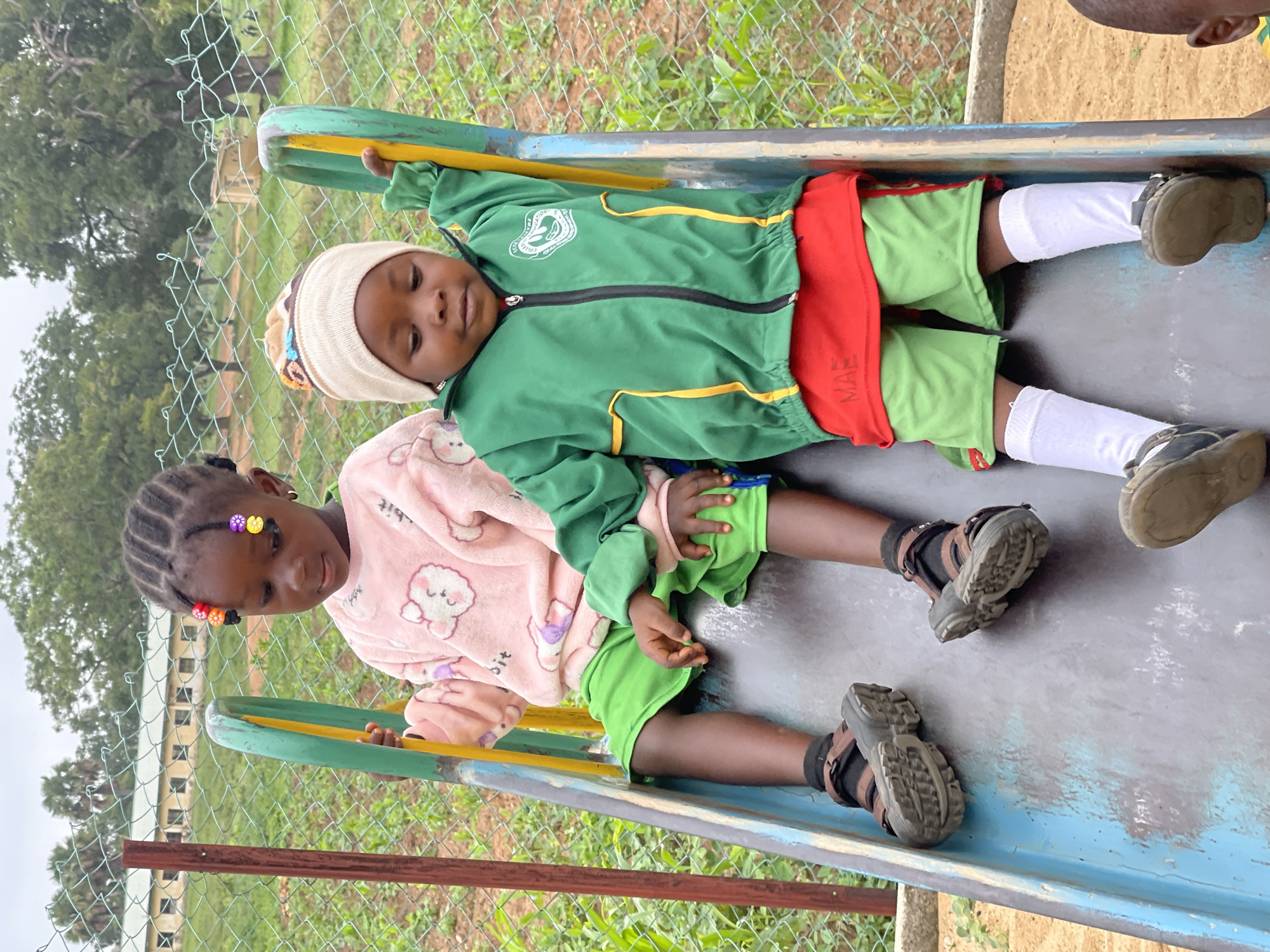 Two young children are captured in a vertical full shot, lying down on a blue-grey slide in an outdoor setting, likely a playground or schoolyard in Akwanga, Nigeria. The image is taken from a slightly elevated perspective, looking down at the children.
The child on the left has dark skin and braided hair adorned with colorful beads. They are wearing a light pink, long-sleeved top with white and purple cartoon sheep motifs, green shorts, and dark sandals with brown straps. This child looks towards the right with a calm and observant expression.
The child on the right has slightly lighter skin and is wearing a cream-colored knit beanie with colorful pom-poms. They are dressed in a green zippered jacket with yellow trim and a white logo on the chest, partially visible as "COLLE..." and an emblem. Beneath the jacket, a red band with the white text "MAE" is visible on their shorts, which are light green. They wear white socks and dark, sturdy shoes. This child looks directly towards the camera with a serene, almost sleepy expression.
The slide they are resting on features green and yellow handrails. In the background, a green chain-link fence separates the play area from an expanse of green foliage and trees. Beyond the greenery, a light yellow building with a brown roof can be seen in the distance, providing a subtle architectural location cue. The ground around the slide appears to be dirt or sand. The lighting is soft and even, suggesting it is daytime, possibly under an overcast sky or during the gentler hours of morning or late afternoon. Both children appear relaxed and content in their positions on the slide.