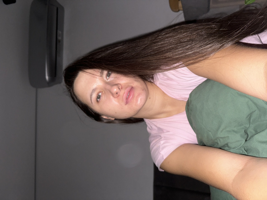 The scene depicts an individual in an indoor setting, identified as being in Molodi, Russia. A single adult female is positioned centrally, facing directly towards the camera. She possesses long, straight dark brown hair, parted on the right side and cascading over her left shoulder and chest. Her eyes are light-colored. She is wearing a light pink, short-sleeved top with a ribbed texture. Her arms are partially visible as she is covered by a dark green blanket or duvet, which is held up to her mid-chest area. The background consists of a plain light grey wall. A silver or light grey wall-mounted air conditioning unit is visible in the upper right quadrant of the image, attached to the wall. To the left, a dark upholstered furniture piece, possibly a headboard or sofa, is partially visible. The lighting is subdued.