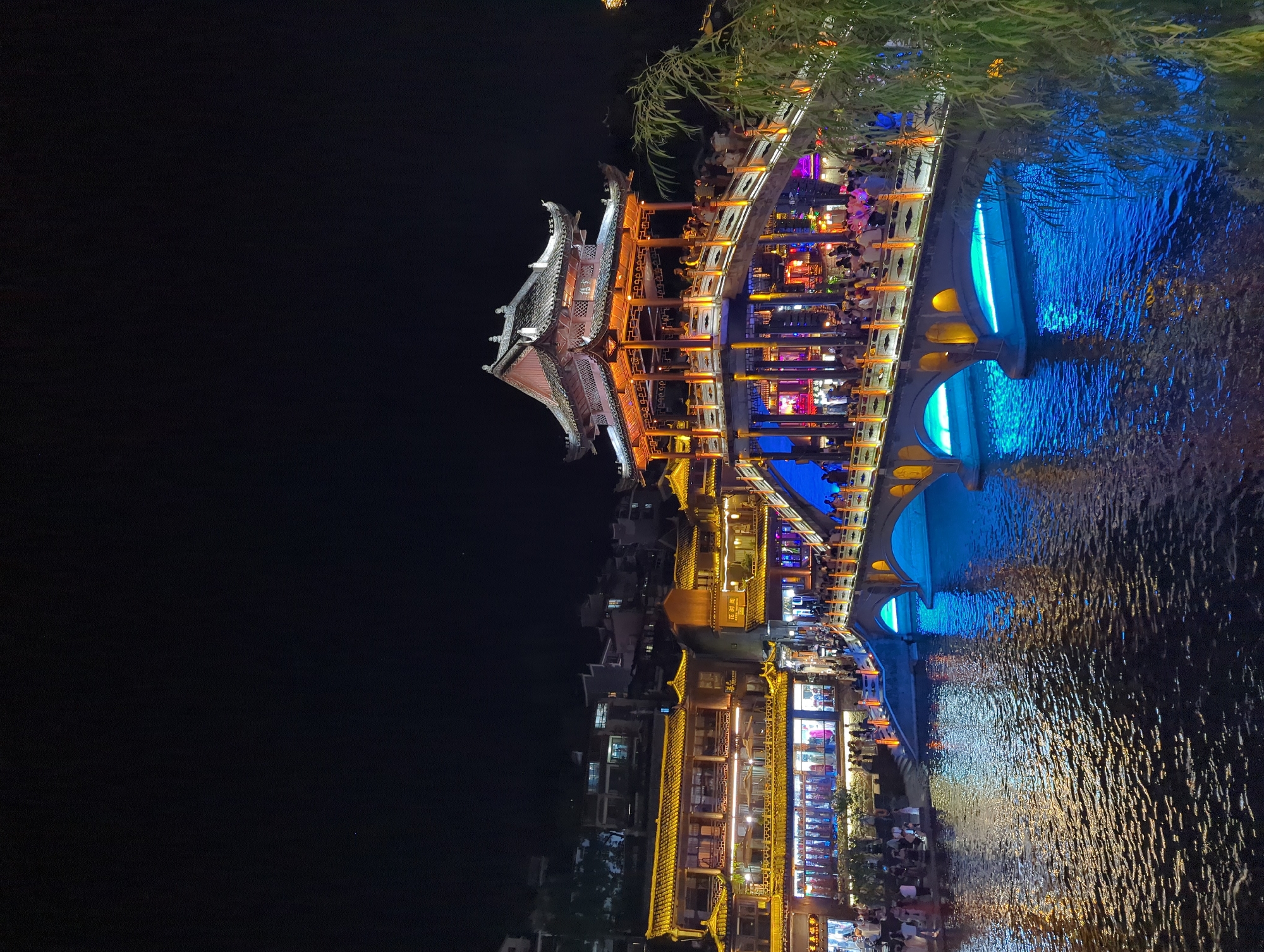 The image depicts the iconic Rainbow Bridge in Fenghuang, China, at night. The bridge, a prominent landmark, is illuminated with warm orange and yellow lights along its railings and structure, highlighting its traditional East Asian architectural style with its multi-tiered roof and intricate carvings. The bridge spans a calm body of water, which reflects the vibrant lights of the bridge and the surrounding buildings, creating a dynamic interplay of colors.
The riverbanks are lined with traditional Chinese buildings, characterized by their distinctive yellow-tiled roofs and ornate facades, also brightly lit with a combination of yellow and white lights. These buildings appear to house shops or restaurants, with visible signs and displays. A significant number of people are present on the bridge and along the riverbanks, suggesting a lively evening atmosphere. They are seen walking, standing, and interacting, many of them casually dressed.
The water itself is a deep, dark blue, interspersed with shimmering reflections of the lights from the bridge and buildings, indicating that it is nighttime and the area is well-lit. The dark night sky serves as a backdrop, accentuating the illuminated structures. In the foreground and to the right, the dark silhouette of a tree with green foliage adds a natural element to the scene, its branches partially obscuring the illuminated bridge. The overall impression is one of a picturesque and bustling tourist destination at night, showcasing the cultural and aesthetic appeal of Fenghuang.