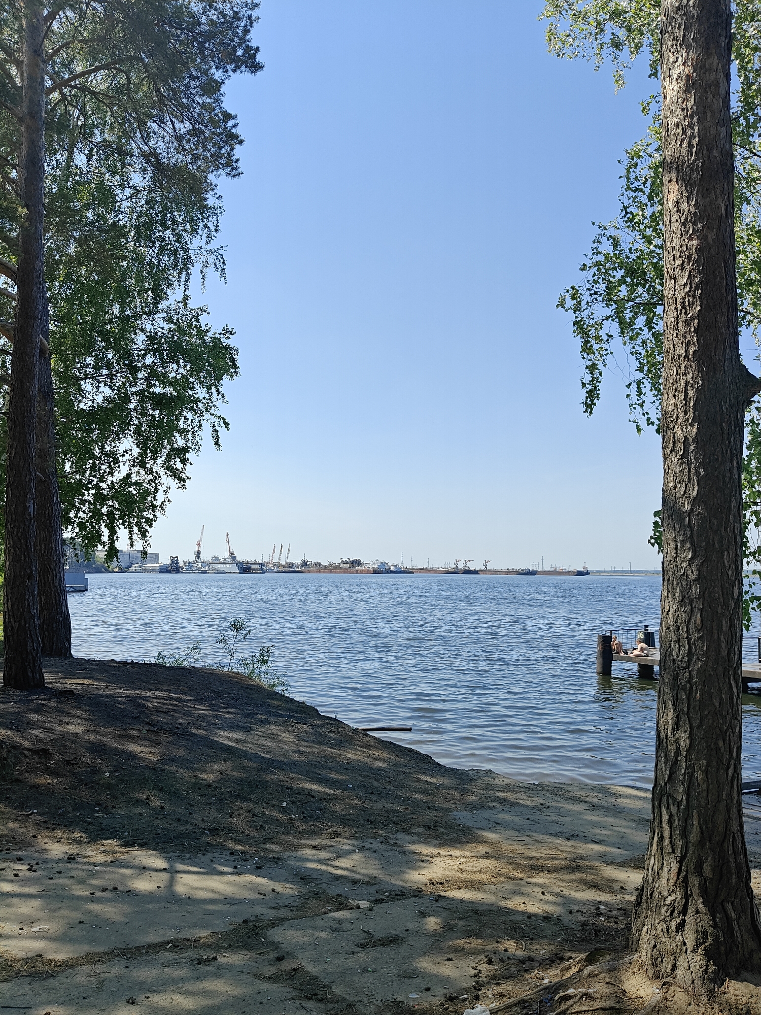 The image depicts a waterfront scene in Chaikovsky, Russia, with a view of the Kama River. The foreground is framed by two trees; one on the left and one on the right, with branches partially obscuring the view. The ground near the trees is covered in dirt and sand, with visible tree shadows. In the middle ground, the Kama River stretches toward the horizon under a clear blue sky. Docking structures and cranes are visible on the opposite riverbank. A small wooden dock with two individuals sunbathing is positioned on the right side of the image, near the water's edge.