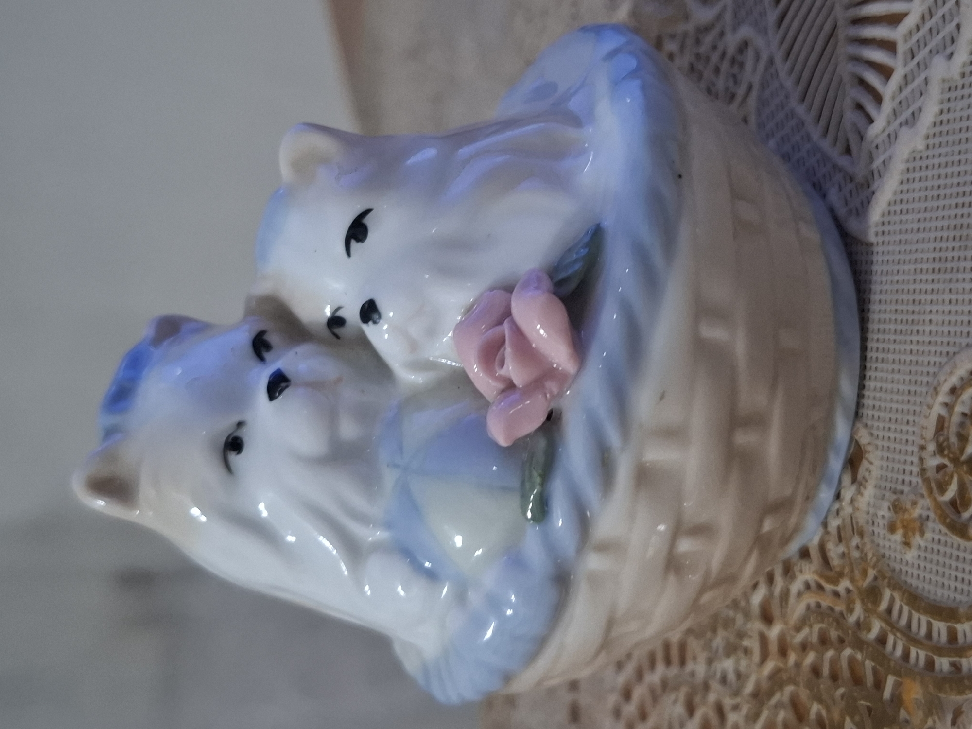 A close-up, eye-level, medium shot captures a ceramic figurine of two white, fluffy cats nestled together in a woven basket. The cats have blue accents on their fur, pointed ears, and black, almond-shaped eyes. A pink rose with green leaves rests beside them in the basket. The basket, with a visible weave pattern, sits on a textured, lacy doily. The background is a soft, out-of-focus blur of neutral tones. The lighting is soft and diffused, highlighting the glossy finish of the ceramic. The scene is indoors, with no specific city or environmental details discernible to place it in Almaty, Kazakhstan.