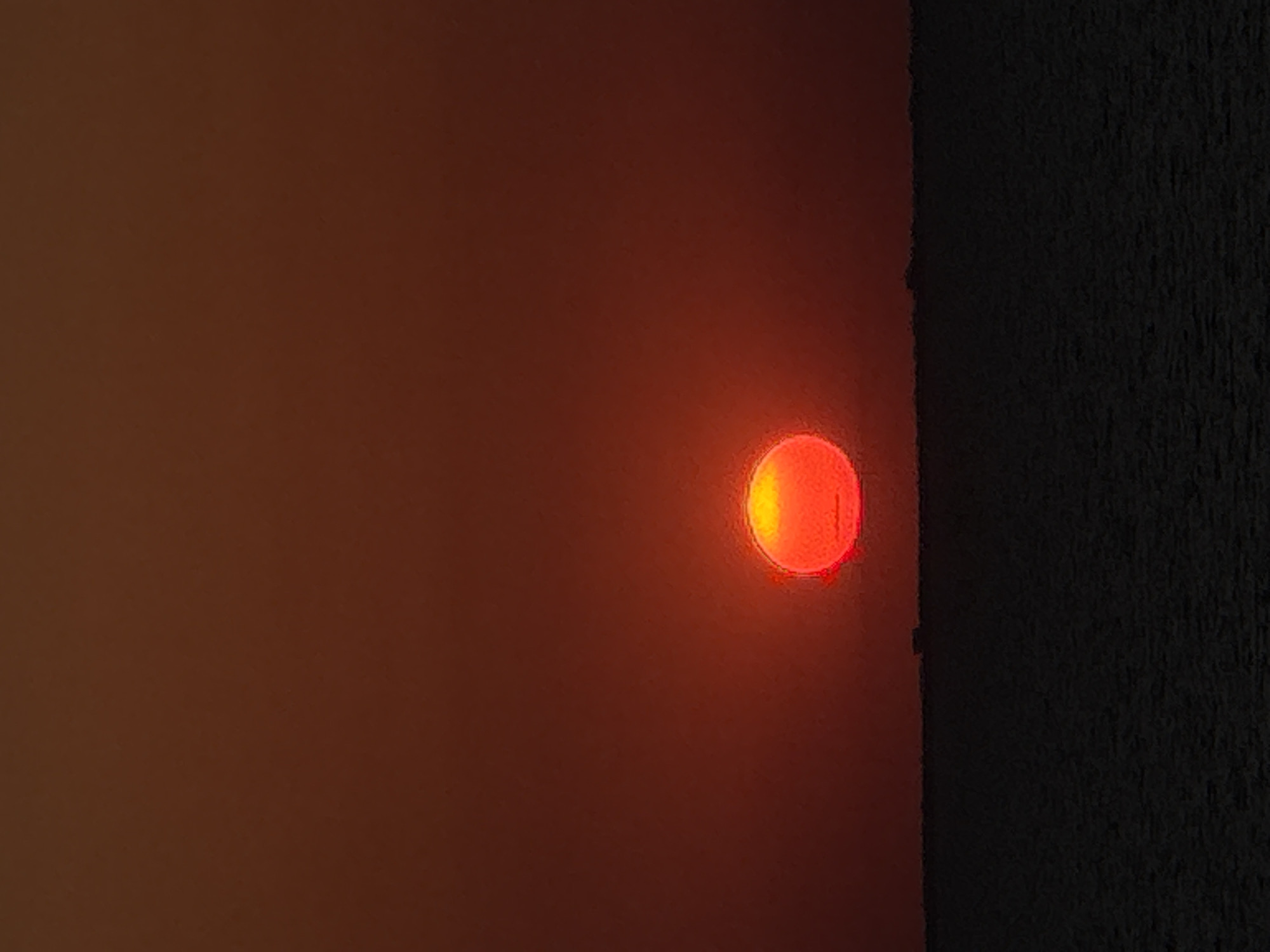 This is a photograph of a red circular light against a dark, textured surface, possibly wood. The light is glowing intensely, with a bright yellow center that fades to red at the edges. It appears to be mounted on a wall or panel. To the left of the light, the surface is a smooth, gradient reddish-brown, becoming darker towards the top left corner. To the right of the light, the surface is a very dark, almost black, material with a pronounced vertical texture. The lighting suggests it might be dusk or night, as the red light is the primary light source in the image. The overall mood is somewhat abstract and minimalist, focusing on the interplay of light and shadow on different textures. There is no visible text in the image. The context provided mentions "Unknown, Italy," but there are no visual cues in the image itself to confirm or deny this location.