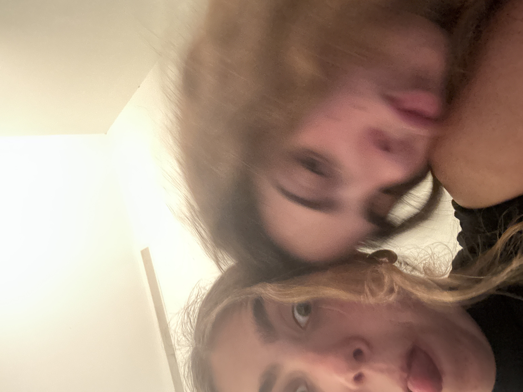 Two young individuals are in close proximity, likely posing for a selfie indoors. The individual in the foreground has blonde hair and is sticking their tongue out. The person behind is blurry with brown hair and also has their tongue out. The background reveals a simple white wall and ceiling corner. There are no indications of Tel-Aviv, Israel.