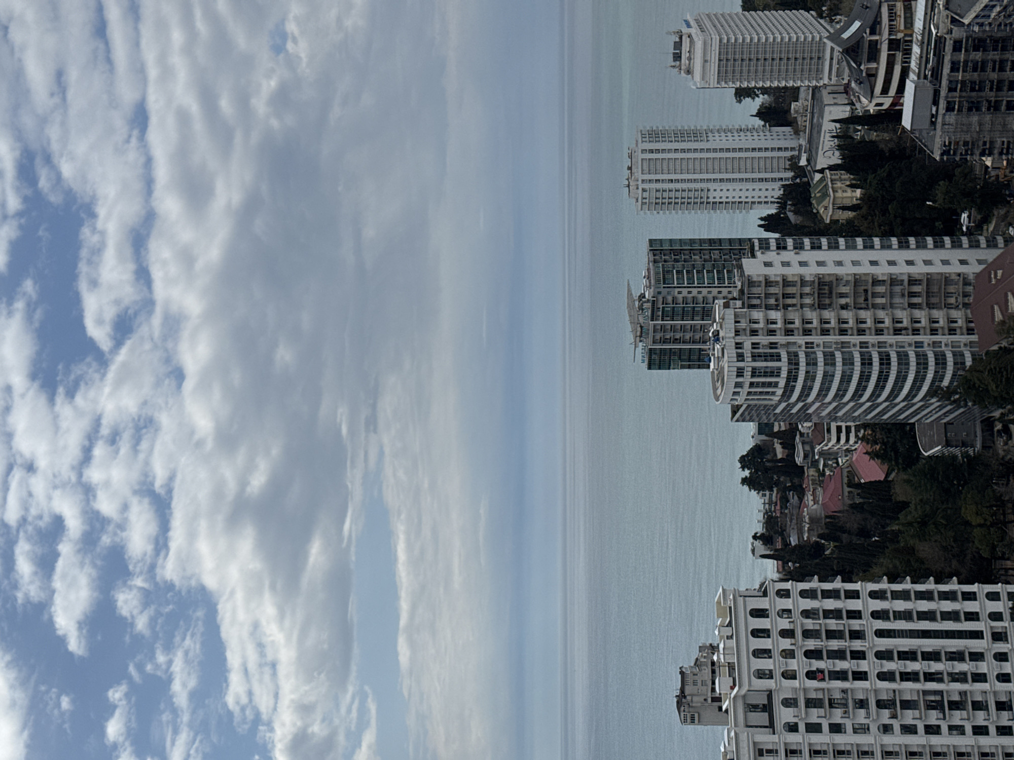 The image shows a view of the city of Sochi, Russia, with the Black Sea in the background. The left side of the image features a sky with scattered clouds. The sea appears calm, with a uniform gray color. The right side shows various buildings of different heights and architectural styles. Some buildings are high-rise structures, while others are lower and nestled among trees. The cityscape includes residential and commercial buildings. The overall atmosphere suggests a modern city by the sea, captured on a cloudy day.