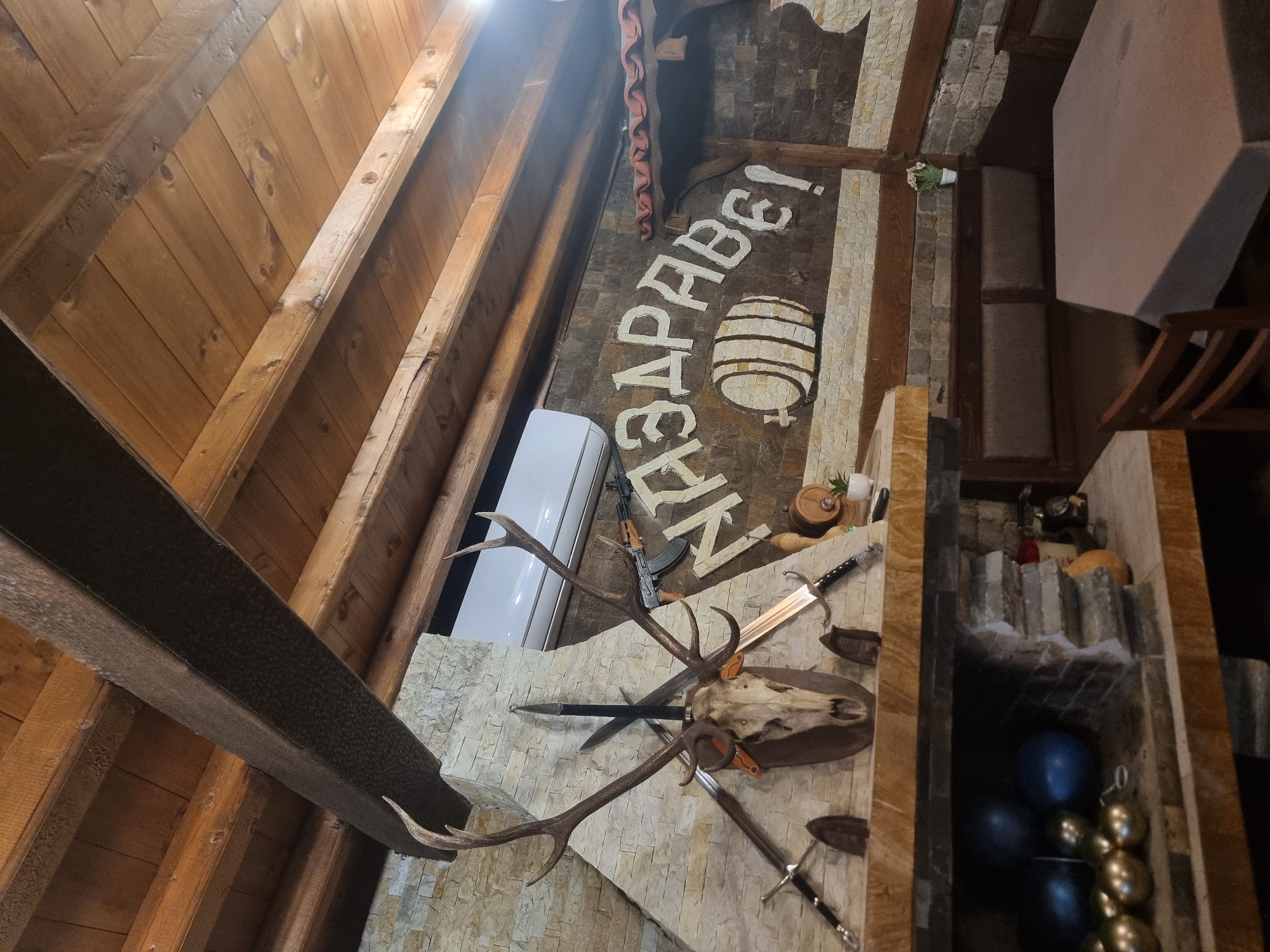 The image shows the interior of a building in Banichan, Bulgaria, possibly a restaurant or bar, judging by the décor. The scene is shot at an upward angle, revealing a wooden beamed ceiling and a stone-clad wall.
On the wall, there is a decorative display including a deer skull with antlers, several swords, and an AK-47 rifle (likely deactivated or a replica). Above this display, there is a painted sign in Cyrillic script, featuring a barrel illustration. A white air conditioning unit is mounted nearby.
To the right of the wall display, there is a glimpse of a seating area with a table and chairs. A decorative niche is also visible, containing blue and gold decorative spheres, along with other objects. The overall impression is a rustic or themed interior design.