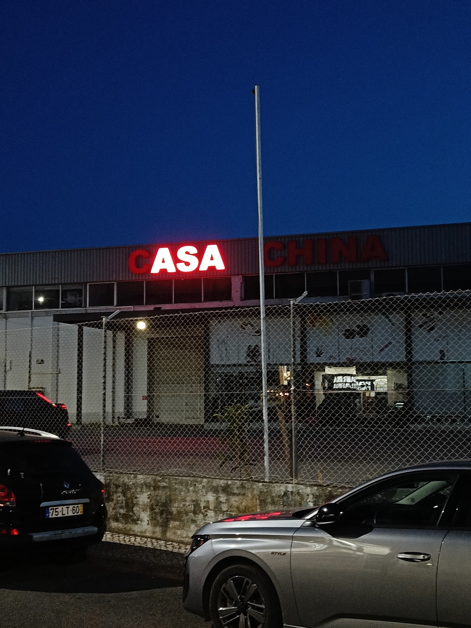 The image depicts a nighttime view of a commercial building with illuminated red lettering on its facade that reads "CASA CHINA". The building is industrial in appearance, with corrugated metal siding and a row of windows. A chain-link fence runs in front of the building, and two cars are parked in the foreground. The sky is a deep blue, suggesting it is dusk or early night. One car, a black SUV, has its license plate visible, reading "75-LT-60". The other car, a silver sedan, is partially visible on the right side of the frame. The overall scene appears to be an exterior shot of a business at night.