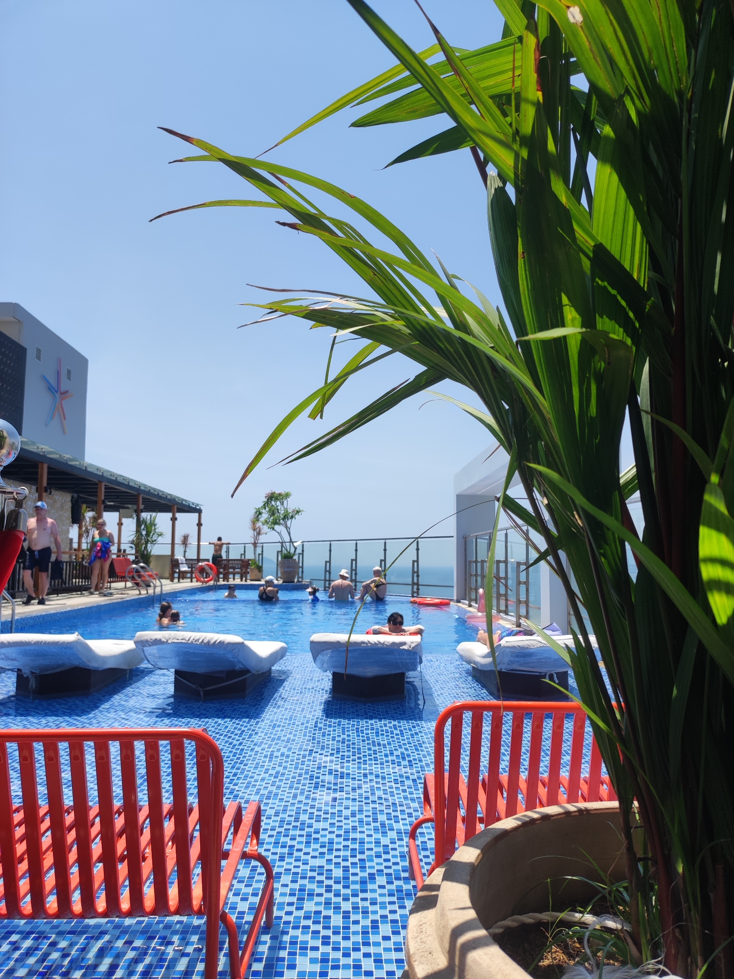The image captures a rooftop swimming pool in Nha Trang, Vietnam, on a clear, sunny day. The pool's surface is a vibrant mosaic of blue tiles. Several people are seen in the water, some swimming and others relaxing. Two individuals are visible on the poolside, one man in shorts and a cap, and a woman in a colorful sarong. In the foreground, a cluster of lush green tropical plants with dark red stems obscures part of the view, adding a natural element to the scene. Beyond the pool, lounge chairs covered in white fabric are submerged in the water, creating a unique seating arrangement. A modern building with a large, star-shaped logo is visible on the left, suggesting it's part of a hotel or resort. The horizon line shows the expanse of the ocean under a clear blue sky. Red outdoor chairs are placed on the tiled deck surrounding the pool.