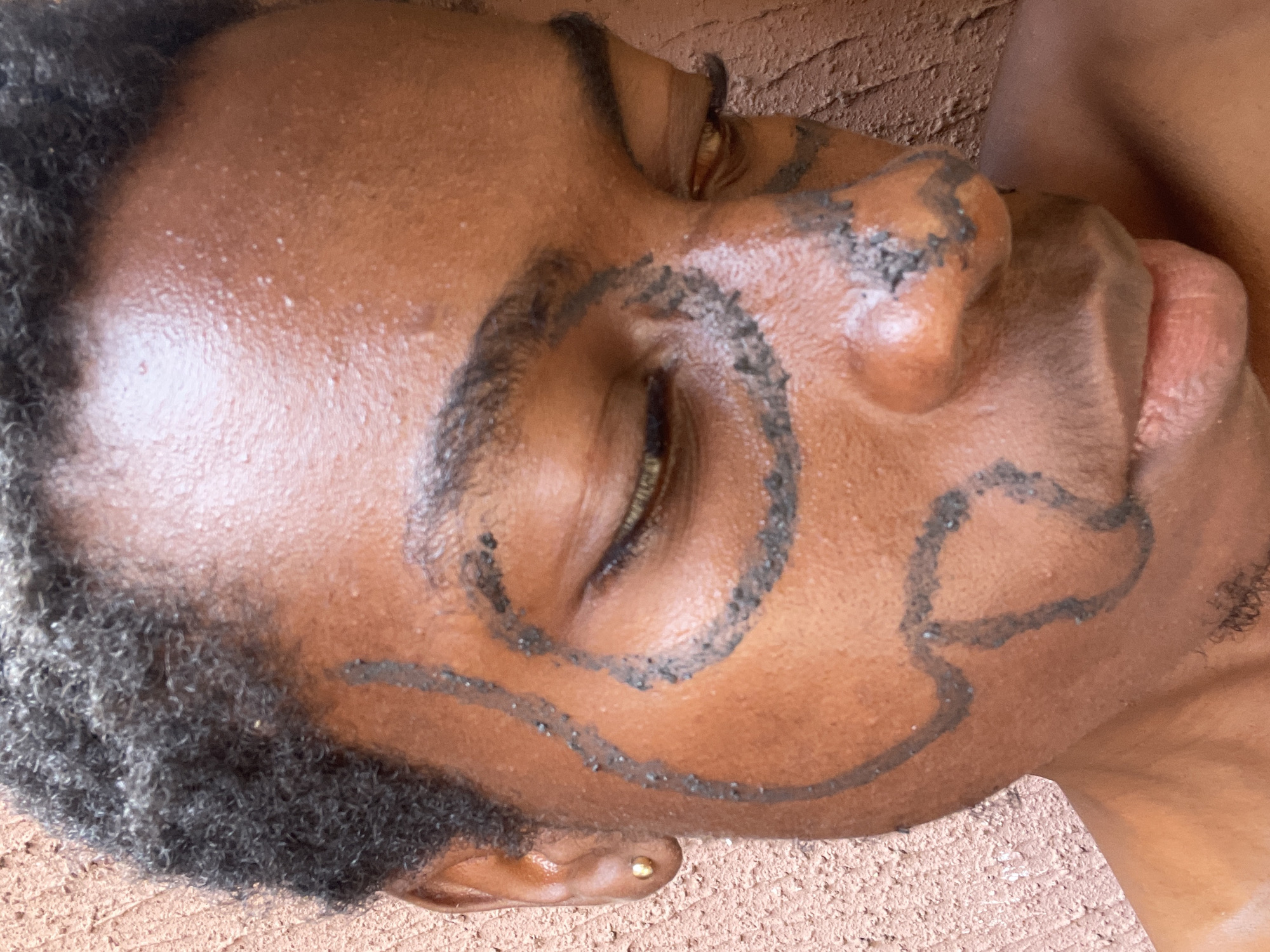 The image is a close-up, vertical portrait of a person's face, seen in profile, with their head resting against a textured, reddish-brown surface. The subject has dark skin, short, tightly curled dark hair, and a small gold stud earring in their visible earlobe.
Their face is adorned with intricate black, granular patterns, possibly created from natural pigments like mud or ash. A prominent circular design encircles the closed eye, and flowing, abstract lines extend across the cheek and jawline, also appearing on the ear. The textured nature of the applied substance is visible, indicating a handmade application. Small white specks are scattered on the skin, particularly on the forehead, which could be dust, dried sweat, or part of the applied material.
The person's eye is closed, conveying a sense of calm or contemplation, and their lips are slightly parted. The background appears to be a wall or ground made of earth or clay, consistent with the location context of Onitsha, Nigeria. The lighting is bright and natural, suggesting it is daytime. No text is visible in the image. The face decoration suggests a cultural, traditional, or artistic event or practice.