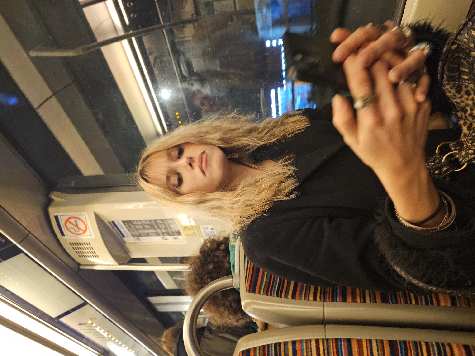A woman with long blonde hair sits on a train. She wears a black coat and several rings and bracelets. The train seat is striped with orange, blue, brown, and yellow. Overhead, there is a "no smoking" sign and a timetable. A window shows a reflection and city lights. The location is Paris, France.