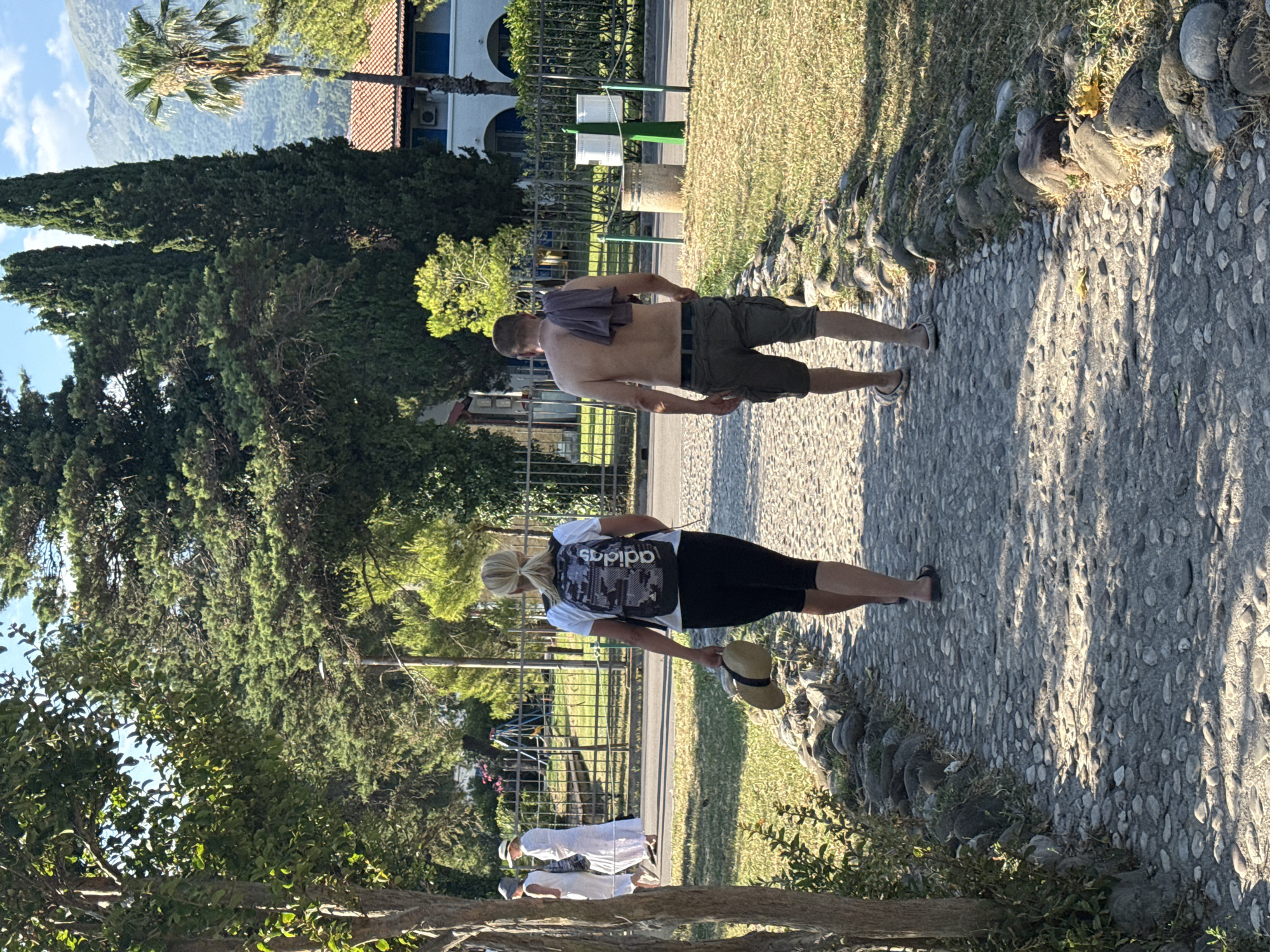 In Budva, Montenegro, two individuals walk along a stone pathway bordering a grassy area. The scene appears to be a park or garden, with lush green trees and a building in the background. The time of day is likely late afternoon, given the long shadows cast by the sun.
The first subject is a man walking with his back to the camera. He is shirtless, wearing dark shorts and has a dark t-shirt draped over his shoulders. He appears to be walking casually.
Further along the path, a woman walks towards the camera, also with her back to the viewer. She is wearing a white t-shirt and black shorts, and carries a backpack with a camouflage pattern that has the "adidas" logo visible. She holds a straw hat in her right hand.
In the background, two other people are visible, dressed in white outfits and hats, suggesting a possibly more formal or traditional attire, or perhaps a tour group.
The setting is outdoors with a focus on the natural landscape and the pathway. The sunlight creates a dappled effect on the ground, indicating a bright, sunny day. The overall atmosphere seems relaxed and leisurely, with people enjoying an outdoor stroll.