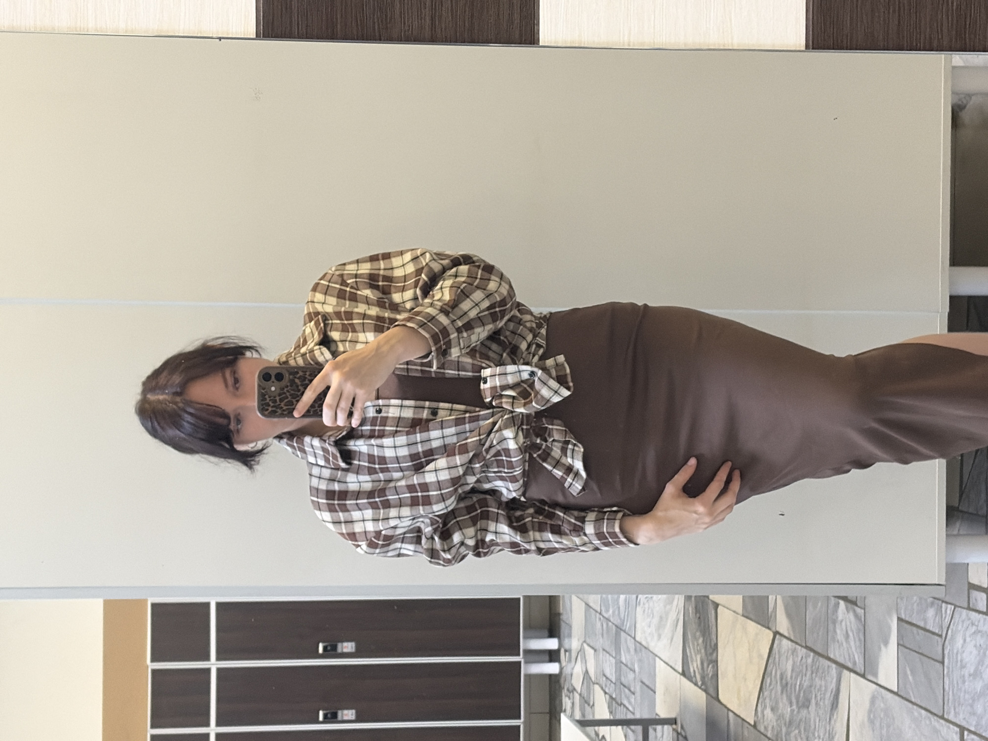 A woman is standing in front of a mirror, taking a selfie. She is wearing a brown and white plaid shirt tied at the waist over a brown slip dress. She is holding a phone with a leopard print case in her right hand. In the background, there are lockers and a tiled floor, suggesting the location might be a gym or locker room. The overall scene is a casual, everyday moment captured through a reflection.